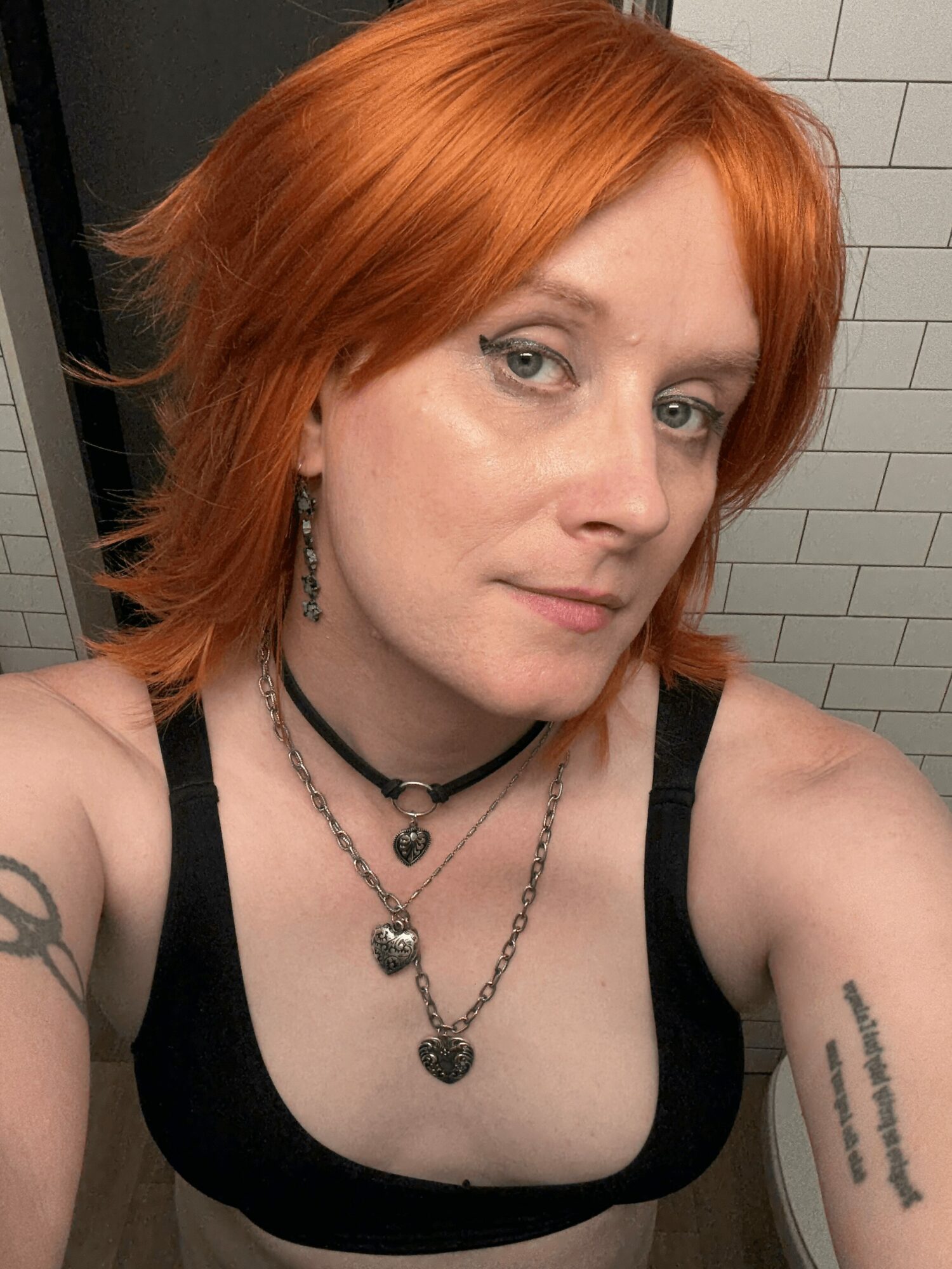 Woman with red hair, wearing black top and layered necklaces, in front of tiled wall.