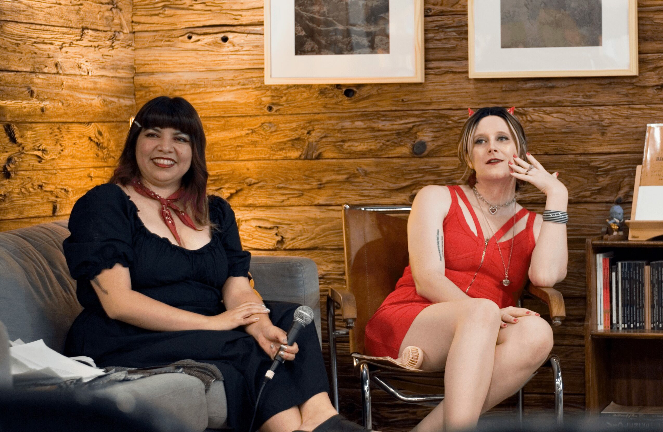 Two women sitting on chairs in a room with wooden walls and framed pictures, one smiling and the other touching her face.