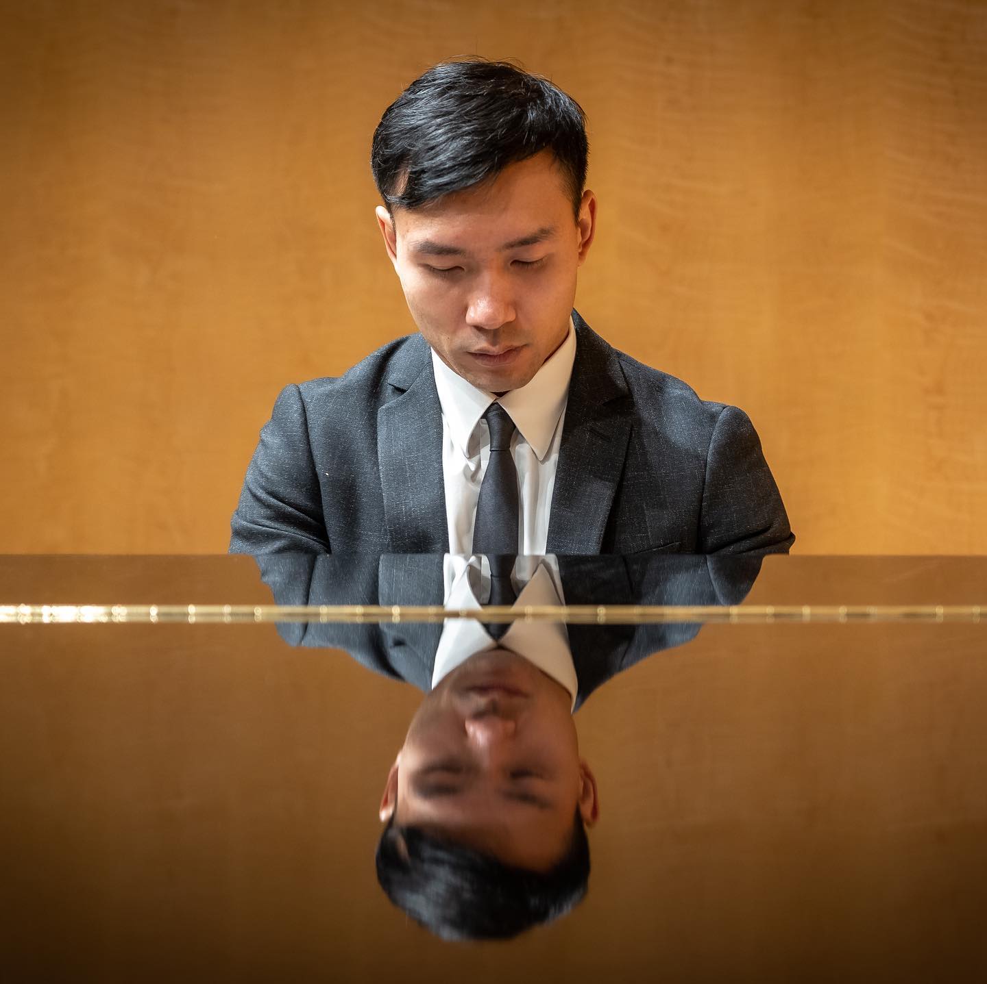 Young man in suit playing piano, reflection visible on shiny surface, warm background, focused expression.
