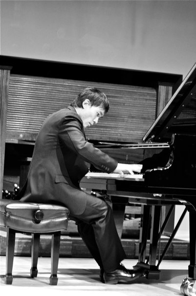 Man playing grand piano, seated on a stool, in a performance setting.