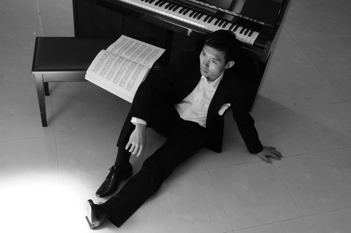 Person in formal attire sitting on the floor next to a piano with sheet music, leaning against the instrument.