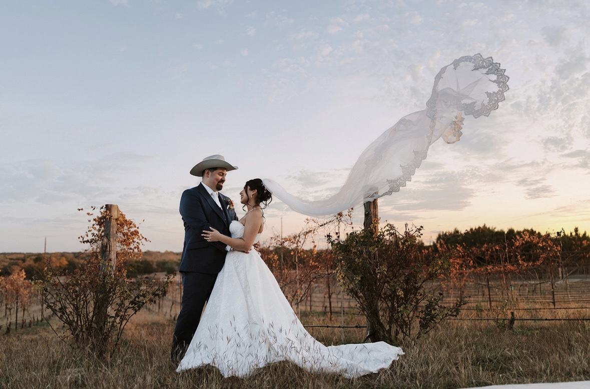 Bride and groom standing outdoors, holding hands, with a large, semi-transparent veil flowing in the sky behind them.