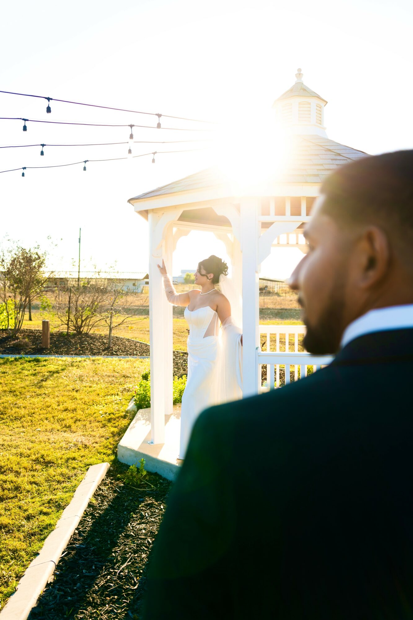 A bride in a white dress and veil stands inside a white gazebo, with sunlight shining behind her outdoors.