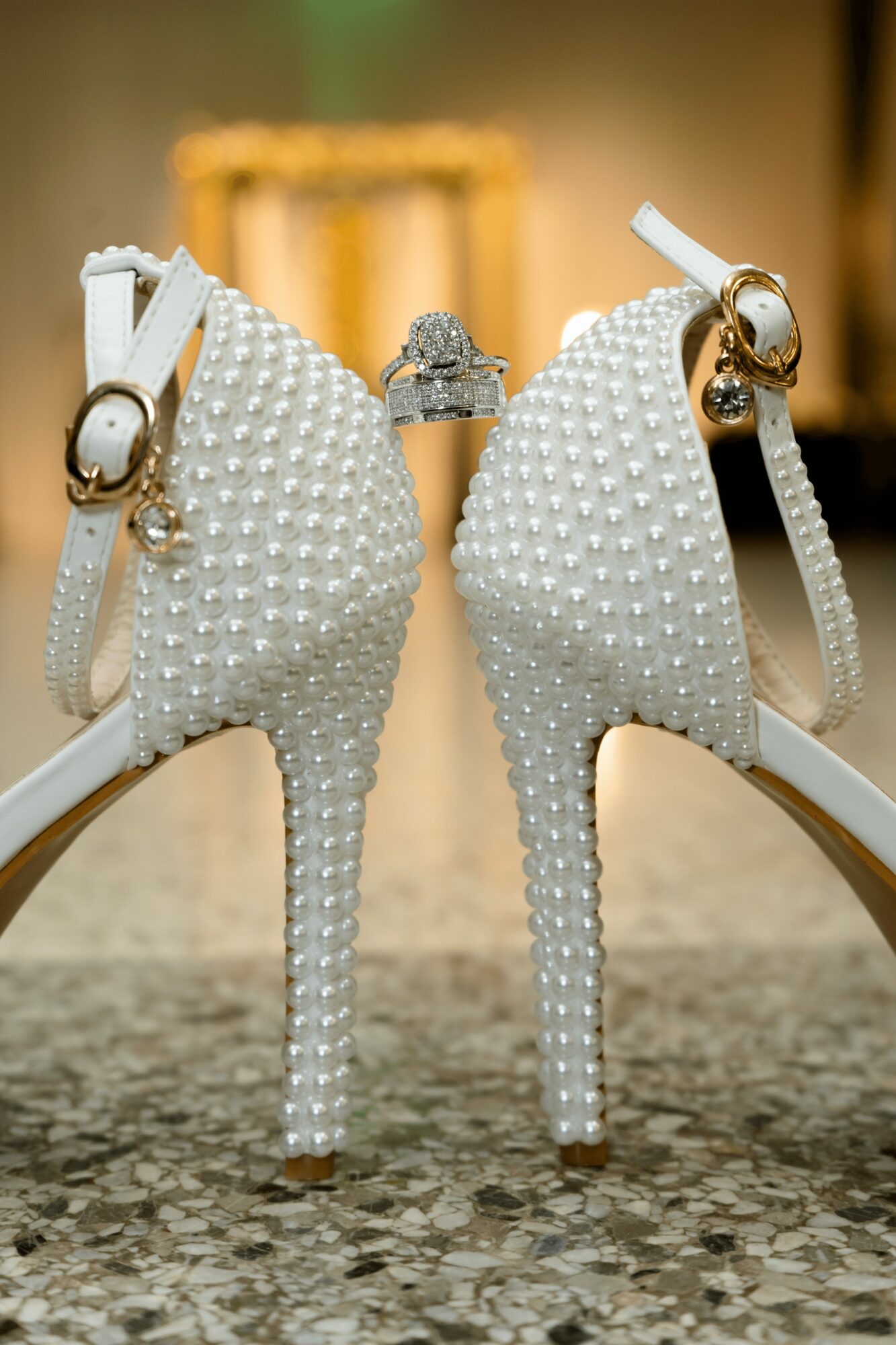 Pair of white high-heeled shoes decorated with pearls, viewed from the back, with rings placed on the heels.