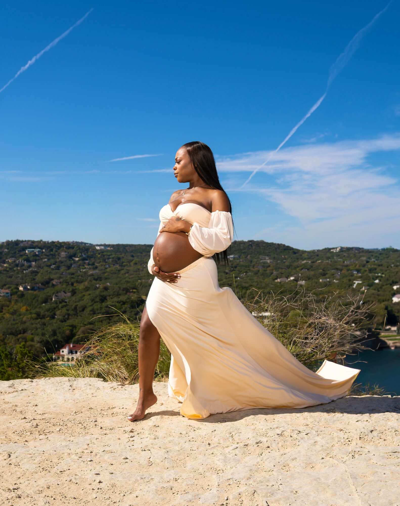Pregnant woman in a flowing dress standing outdoors on a sunny day with a landscape background.