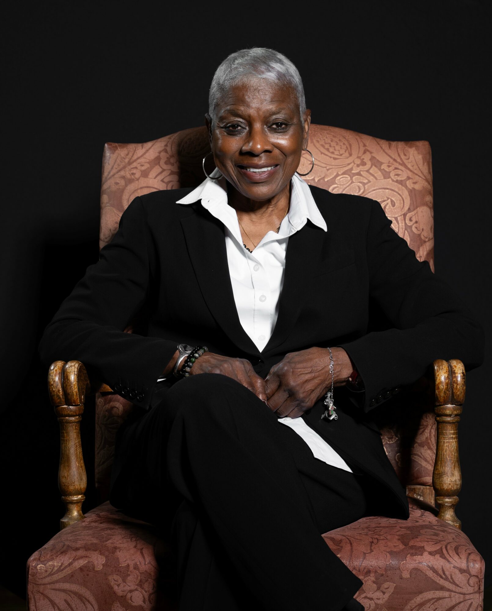 Older woman with short gray hair sitting in a patterned armchair, wearing a black suit and white shirt, smiling.