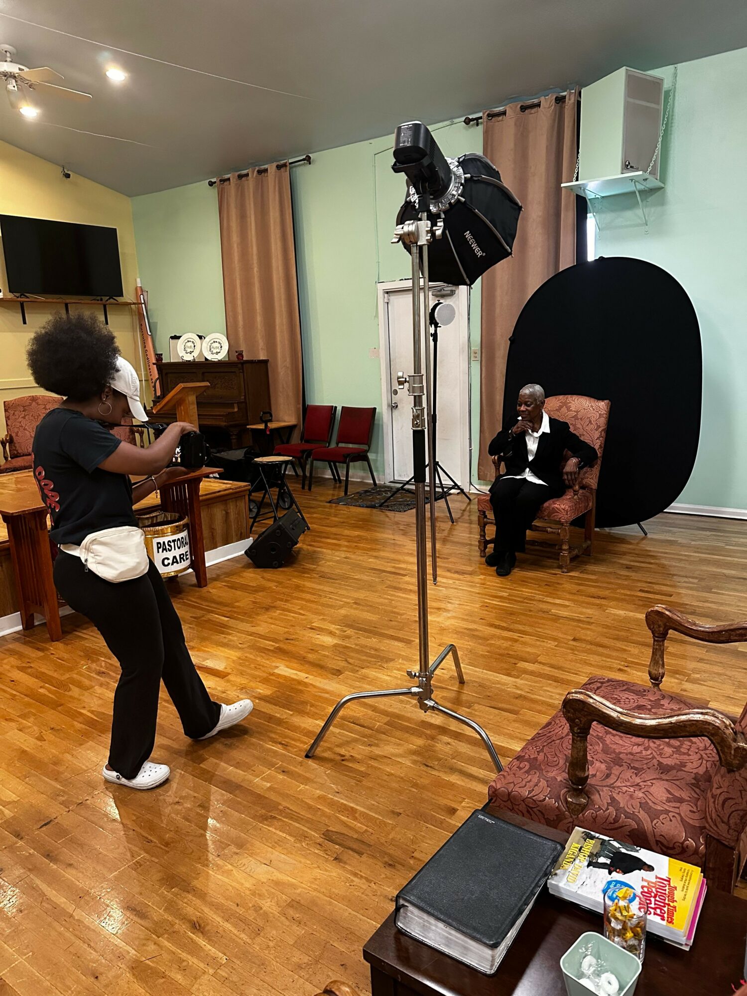 Person standing and holding a camera in a room with a person sitting on a chair, photography equipment, and wooden furniture.