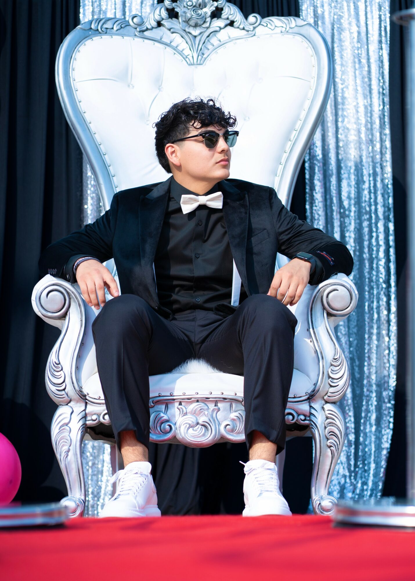 Person sitting on a large ornate white throne, wearing sunglasses, black suit, and white sneakers, with a blue curtain background.