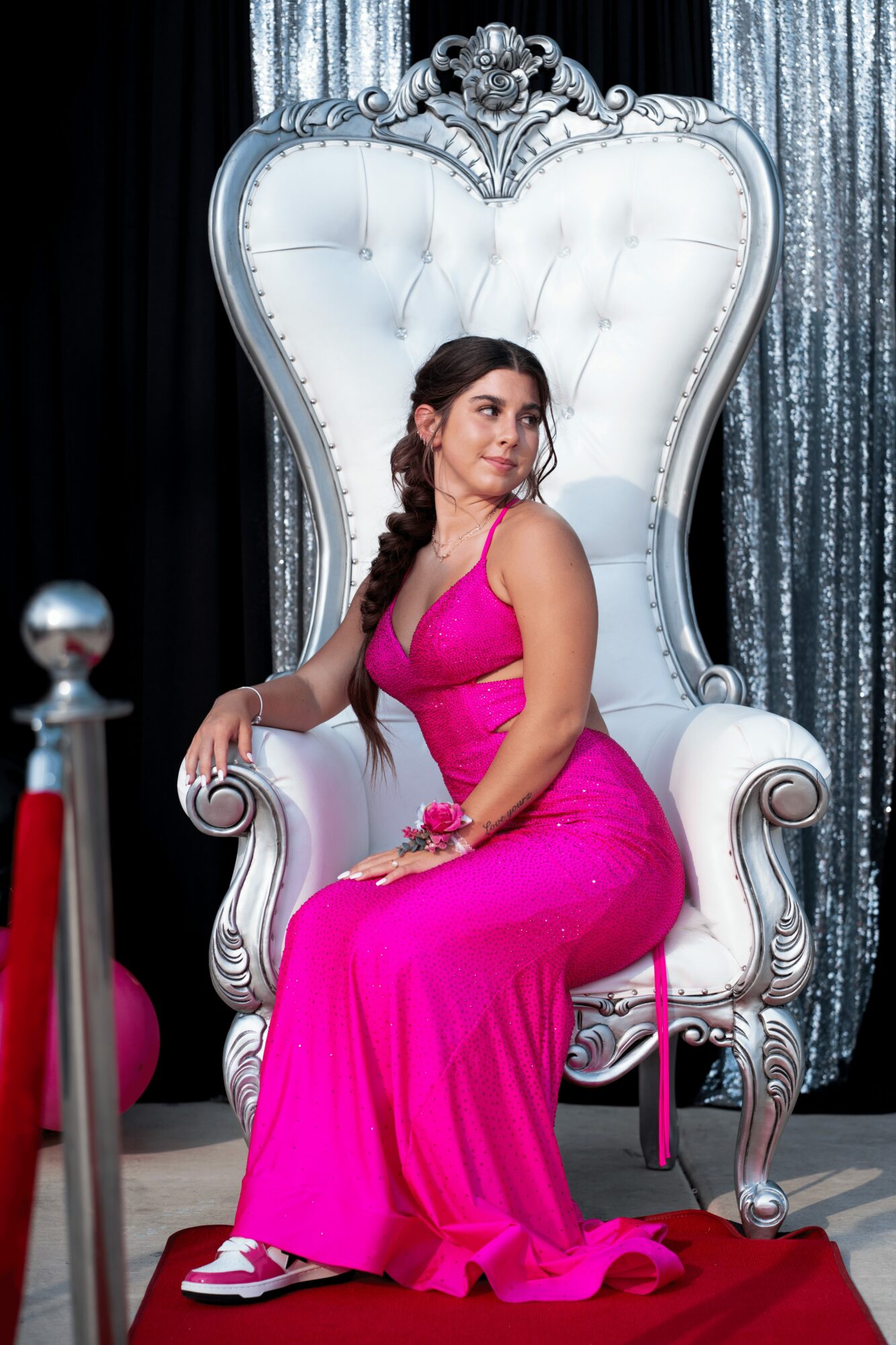 Young woman in pink dress sitting on ornate white throne with silver details, backdrop of black and silver curtains.