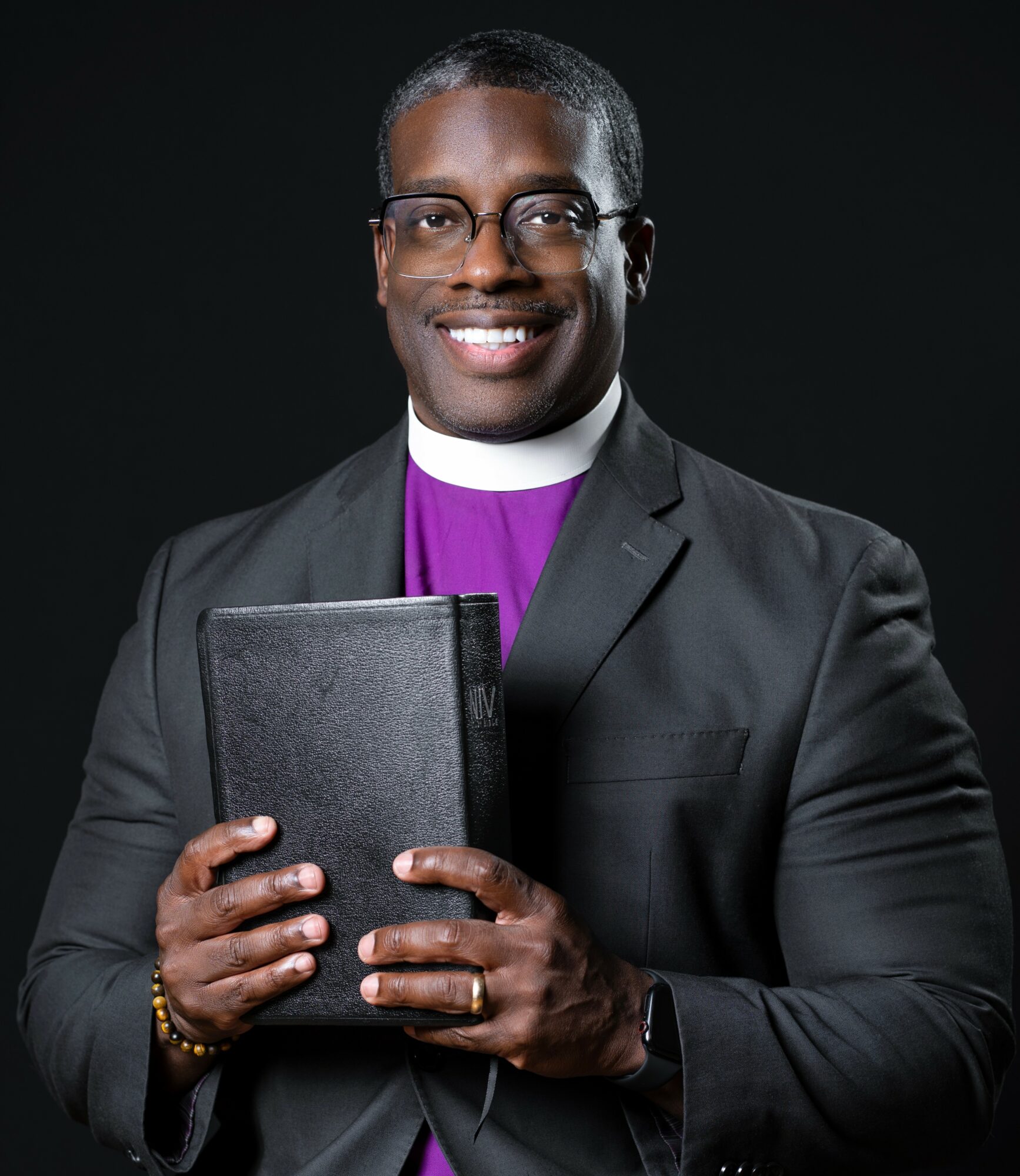 Clergy member in suit holding a black book, smiling, against a dark background.