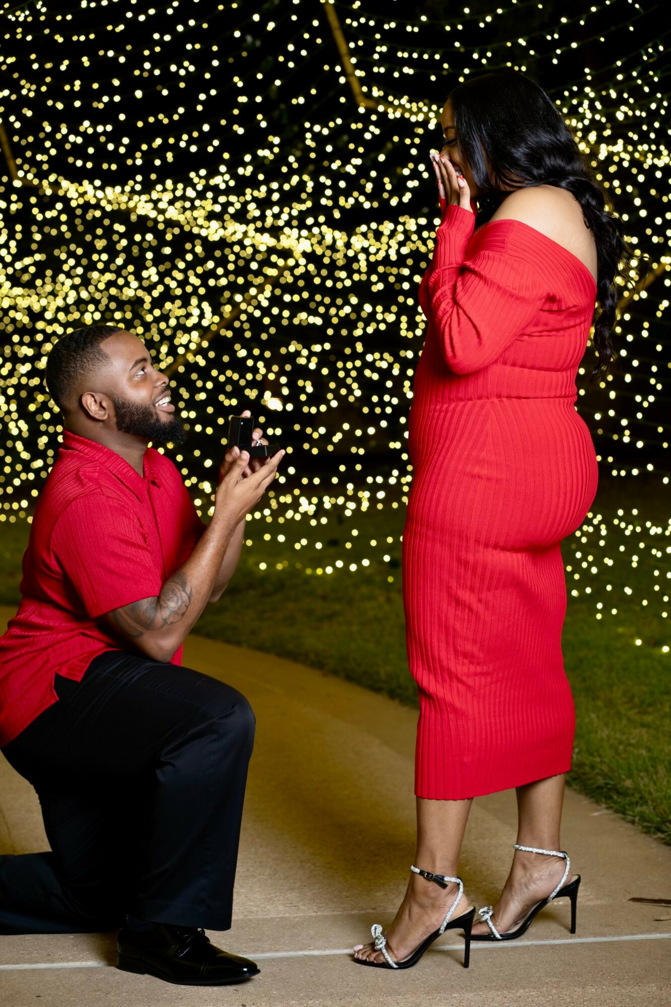 A man kneeling proposing to a woman in a red dress outdoors at night with string lights in the background.