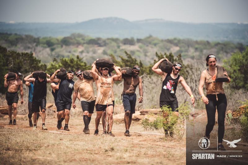 Group of people walking outdoors on a dirt path, some shielding eyes from the sun, with trees and hills in background.