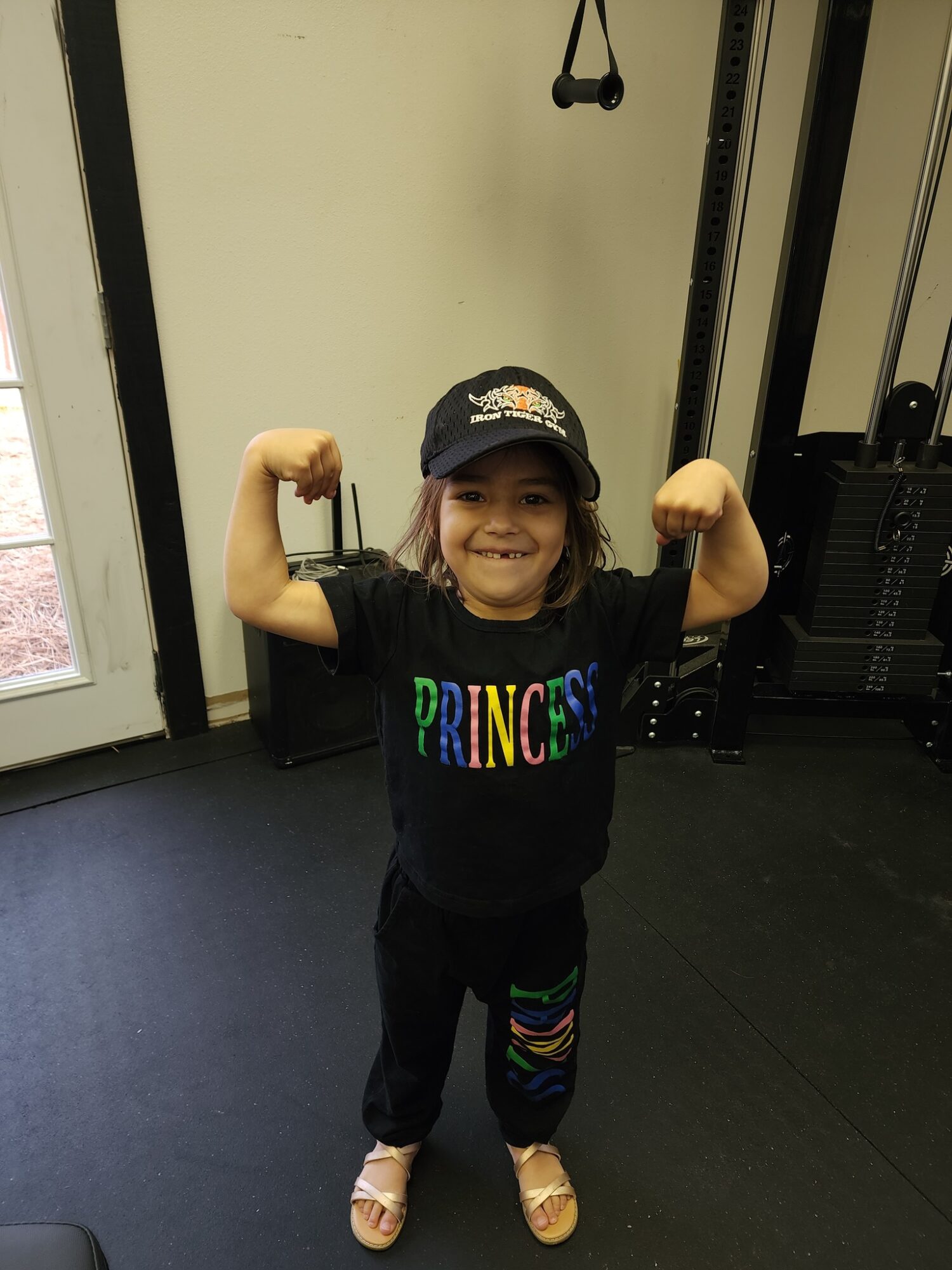 Young girl smiling and flexing her arms in a gym, wearing a black cap and a black shirt with colorful text.