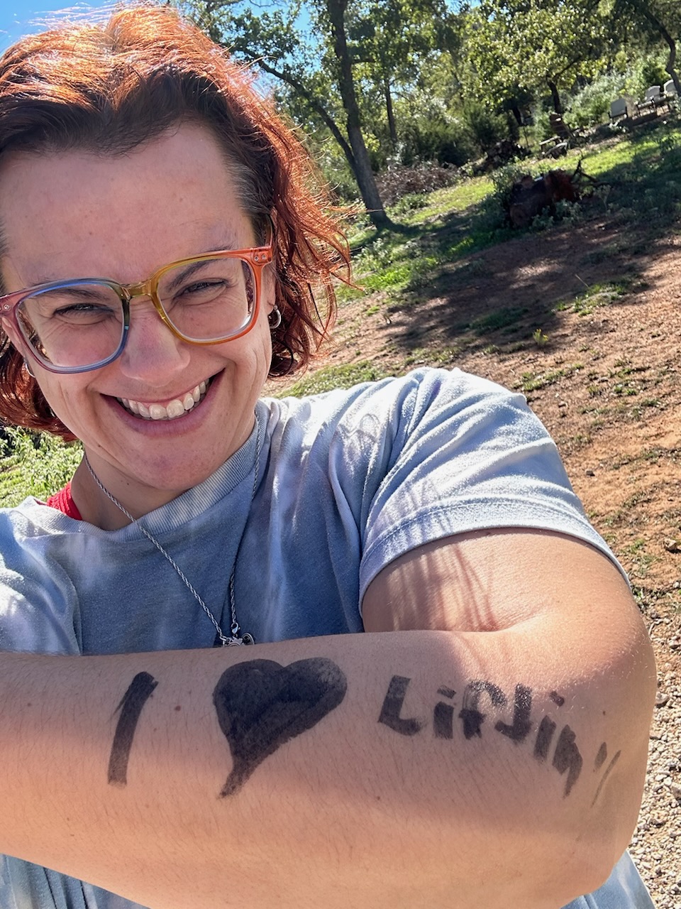 Smiling woman with glasses outdoors, showing a message on her arm with a heart symbol and the name 'Lindsey'.
