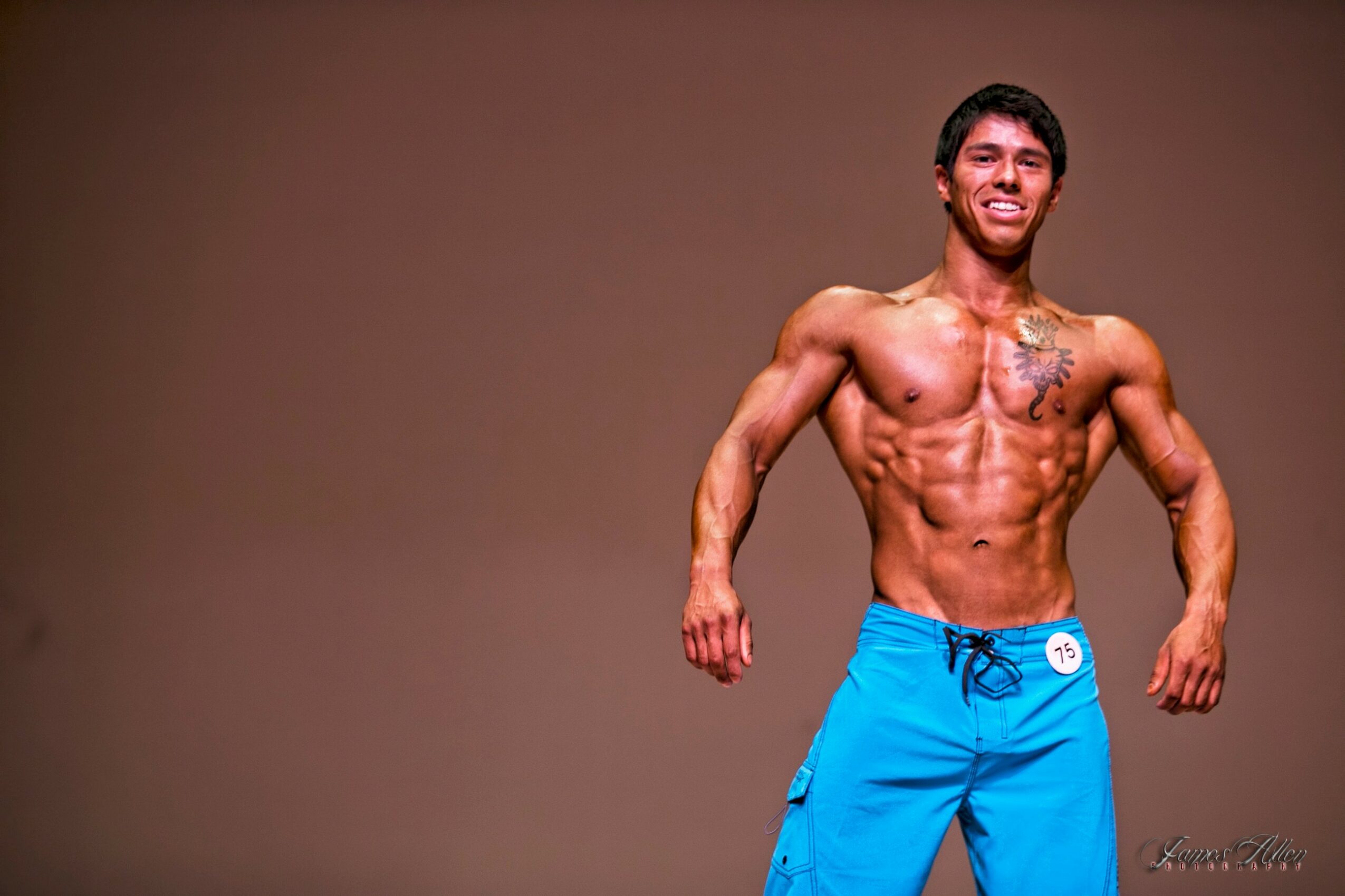 Young man with muscular physique smiling, wearing blue shorts, standing against a plain background.
