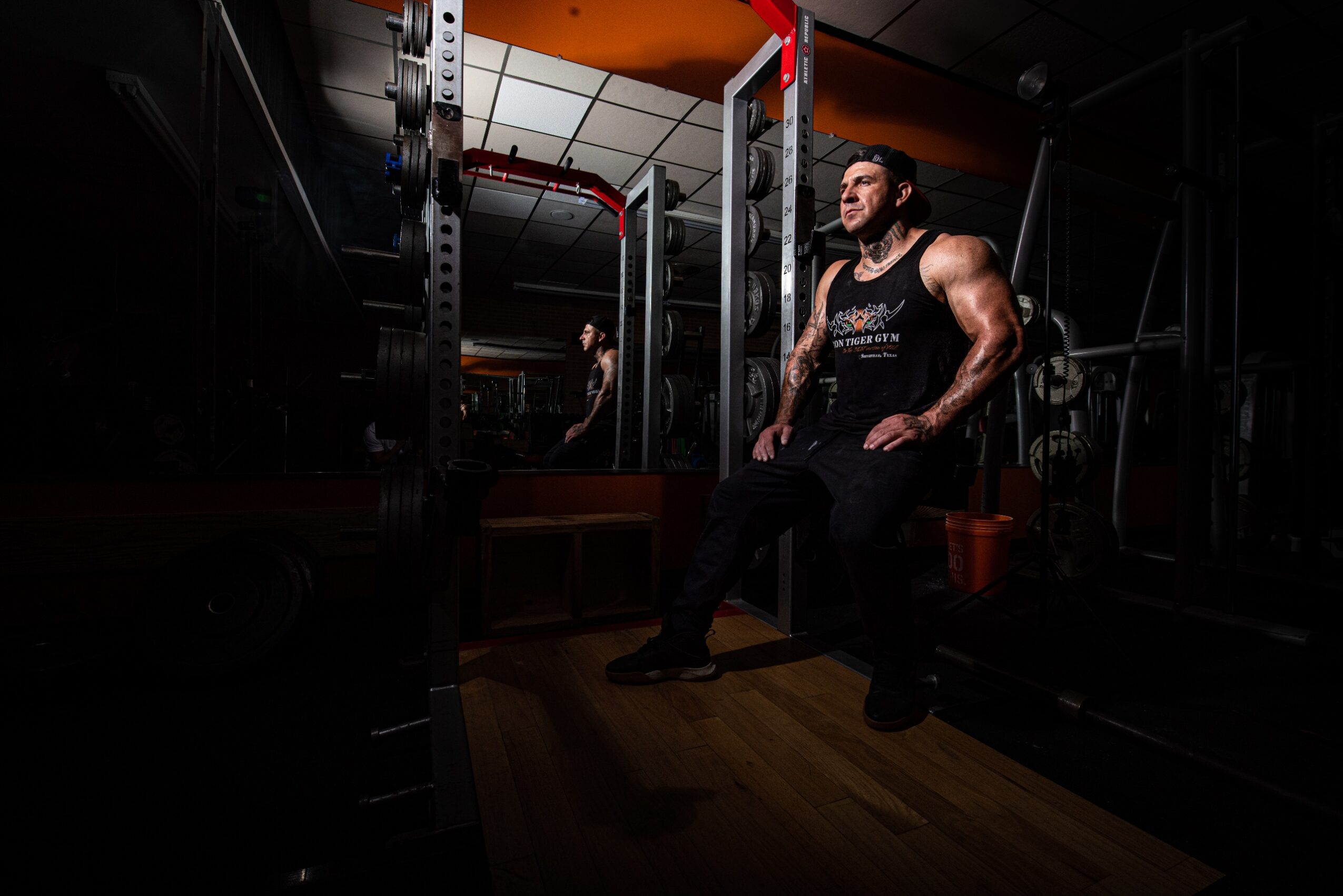 Man sitting on a bench in a gym, surrounded by weightlifting equipment, with a mirror reflecting his image.