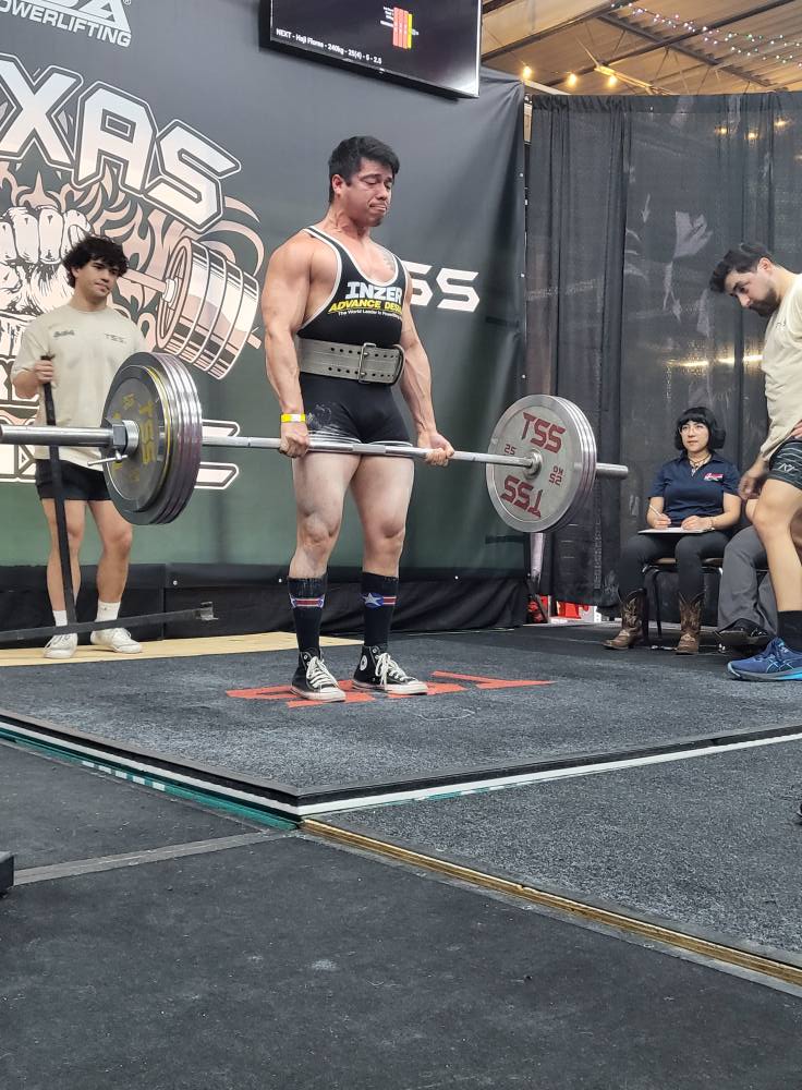 Powerlifter preparing to lift a barbell with weights in a competition setting, with spectators and a banner in the background.