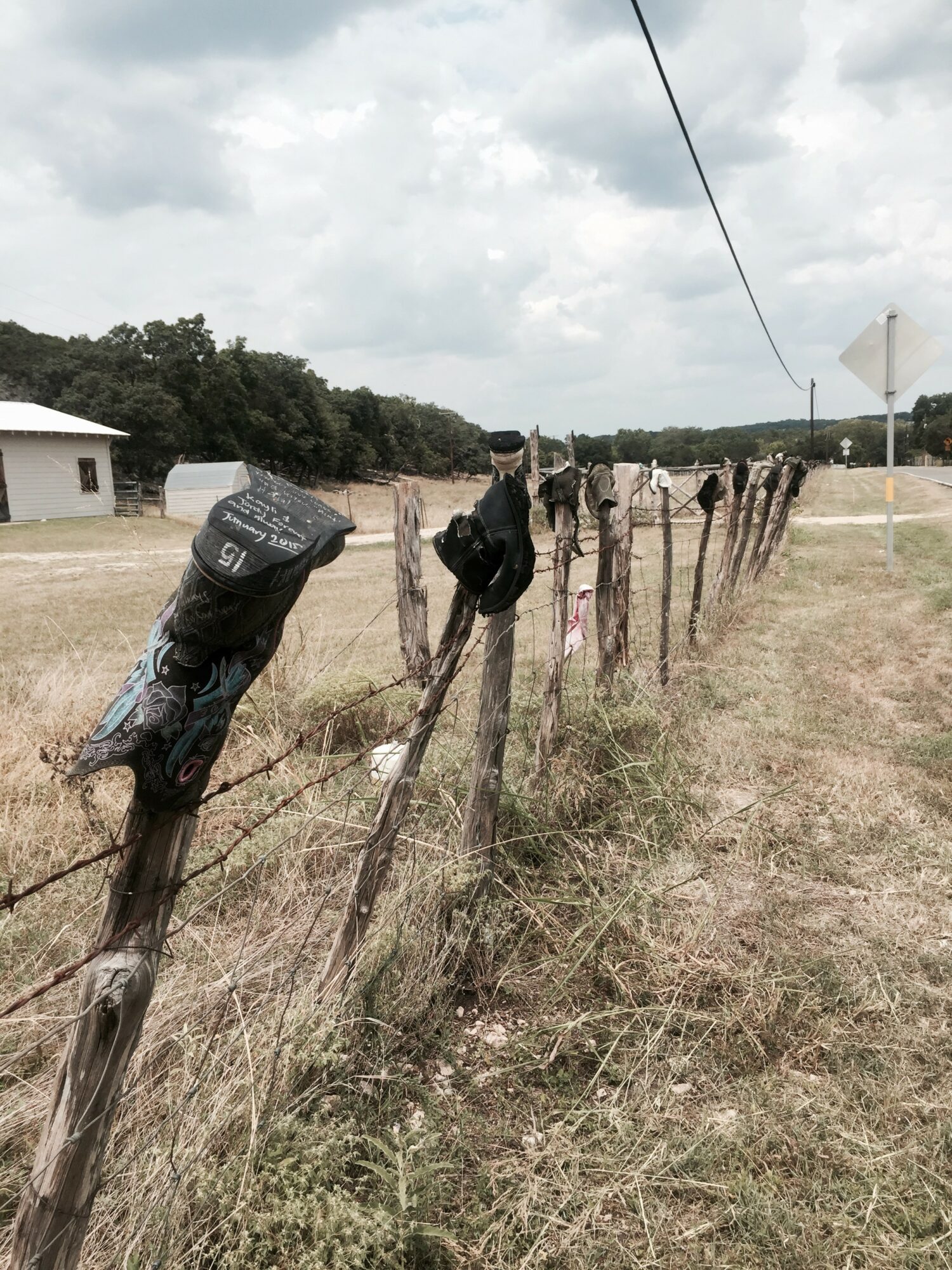 Line of weathered wooden fence posts with backpacks hanging on some, in a rural landscape with cloudy sky and trees.