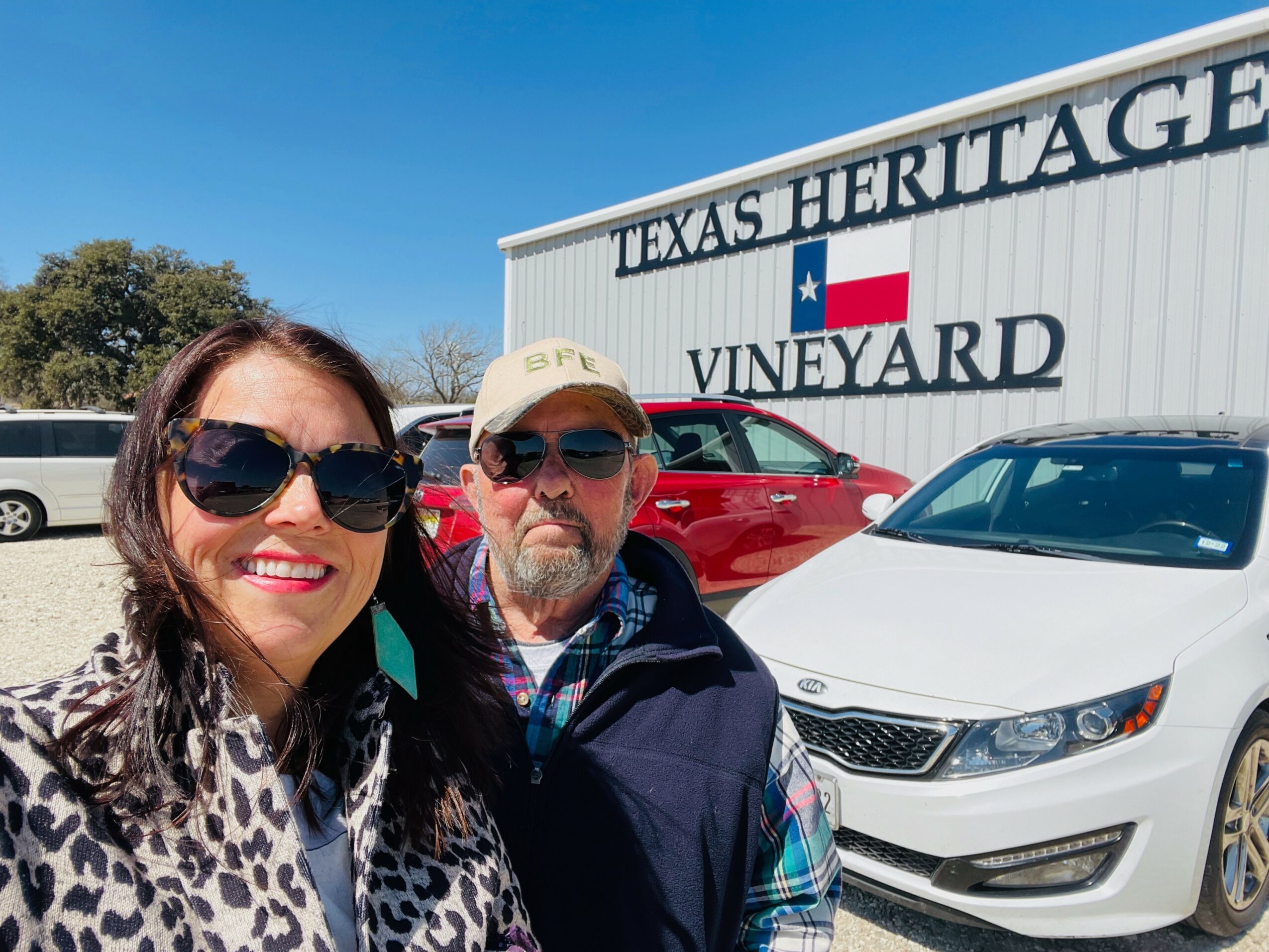 Two people smiling in front of a building with a sign reading 'Texas Heritage Vineyard,' with cars parked behind them.