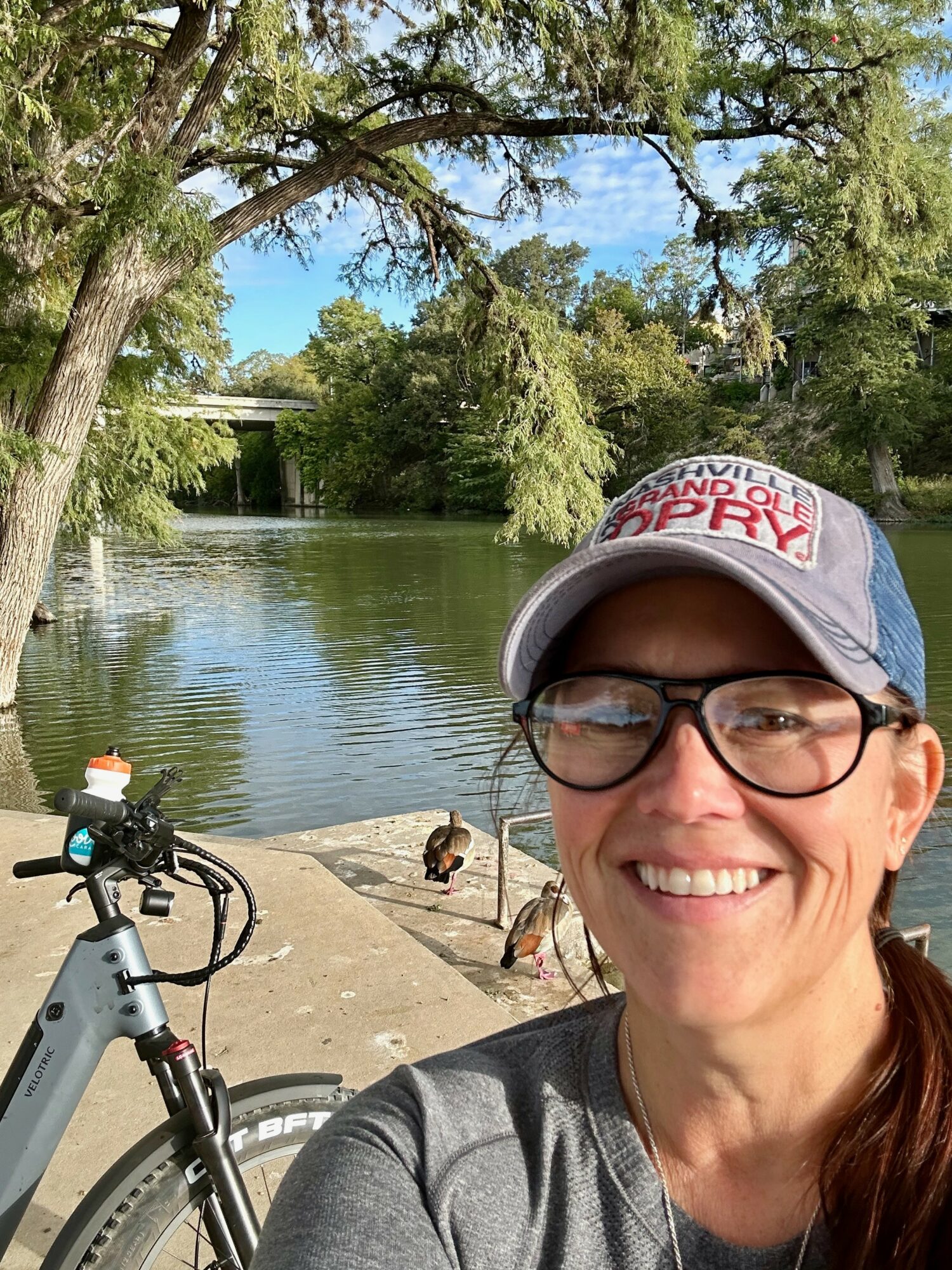 Woman wearing glasses and a cap smiling outdoors near a river with trees and ducks.