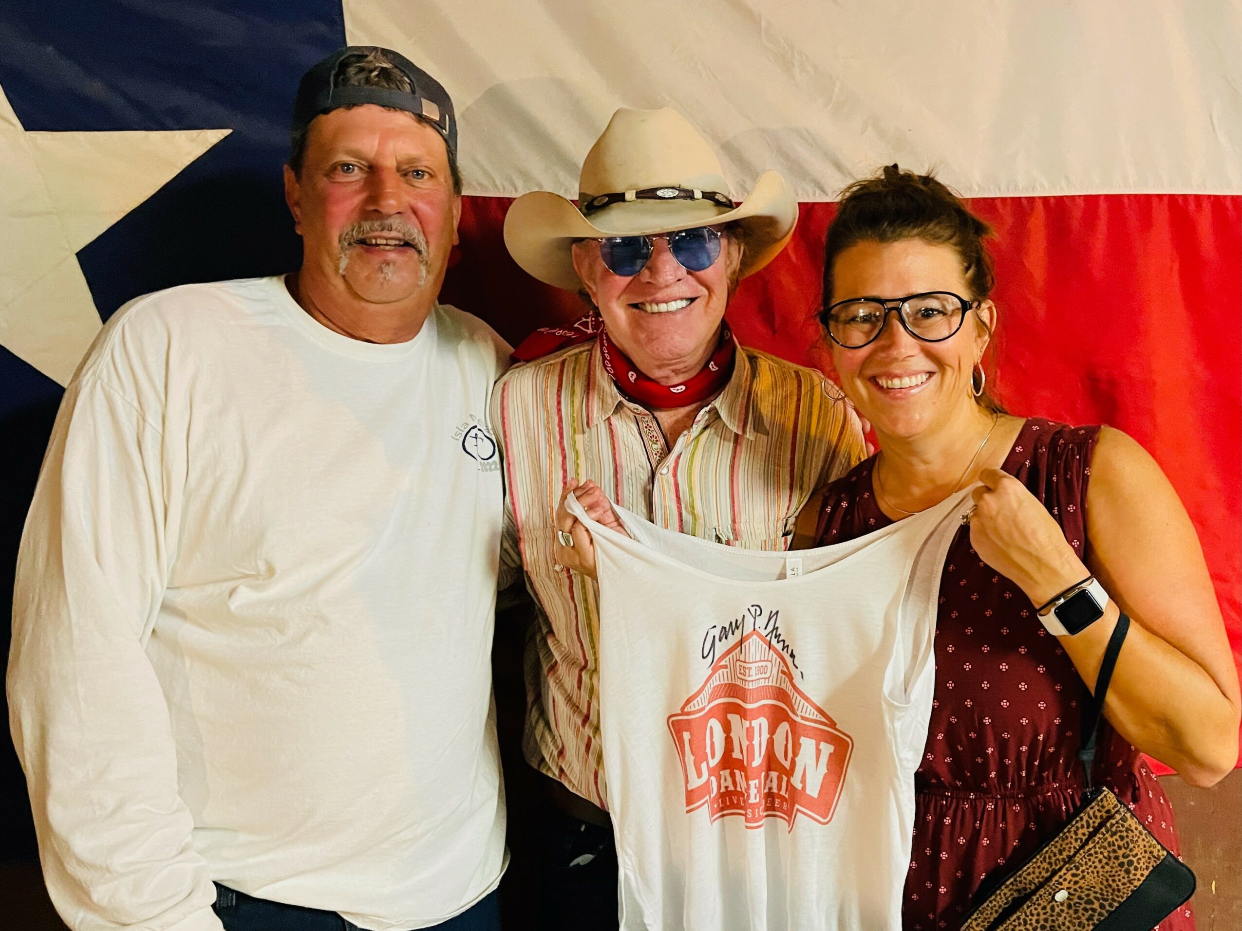 Three people smiling in front of a Texas flag, one holding a white cloth with red text.