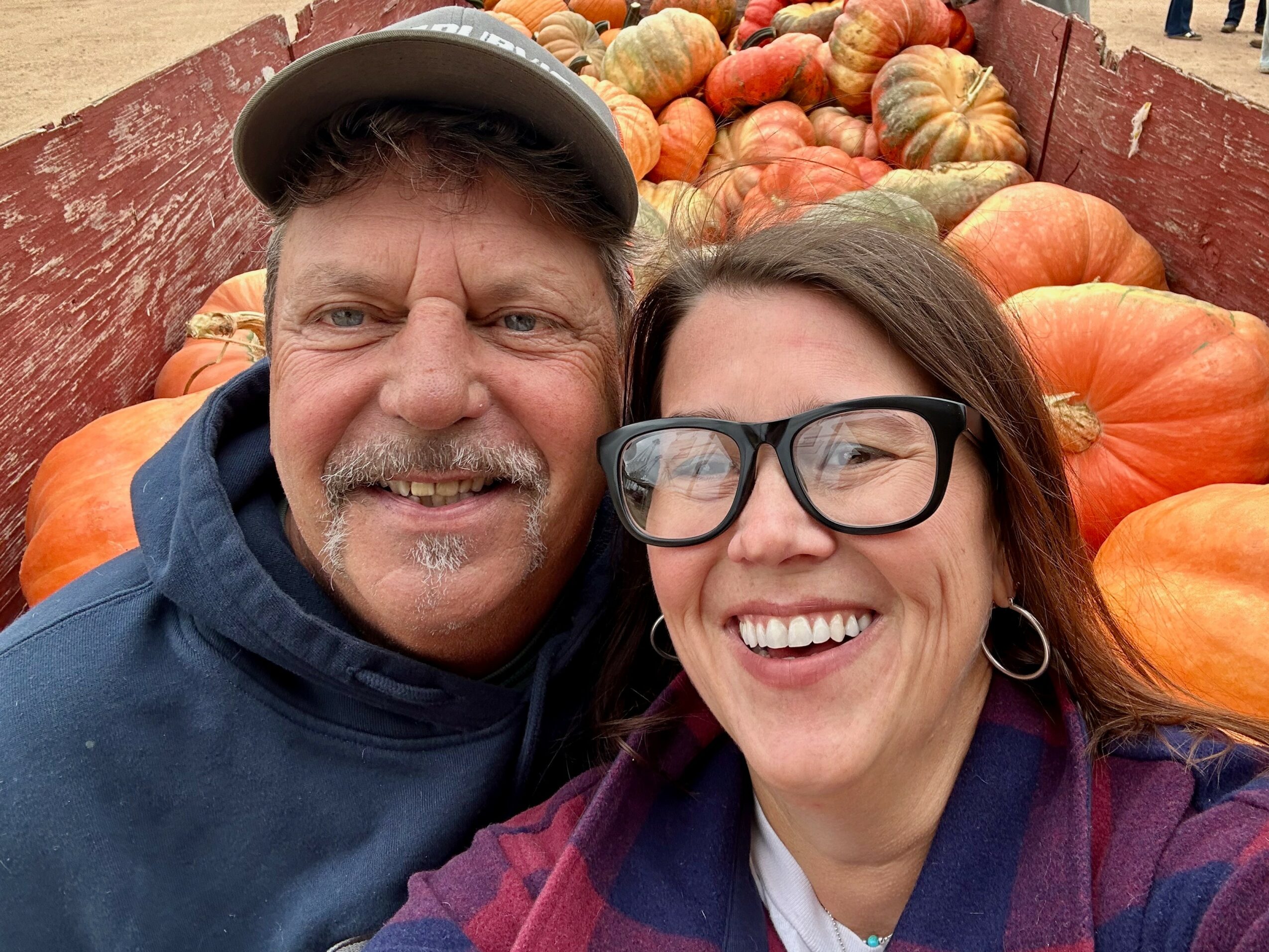 Two people smiling in front of a pumpkin display at a market or farm stand.