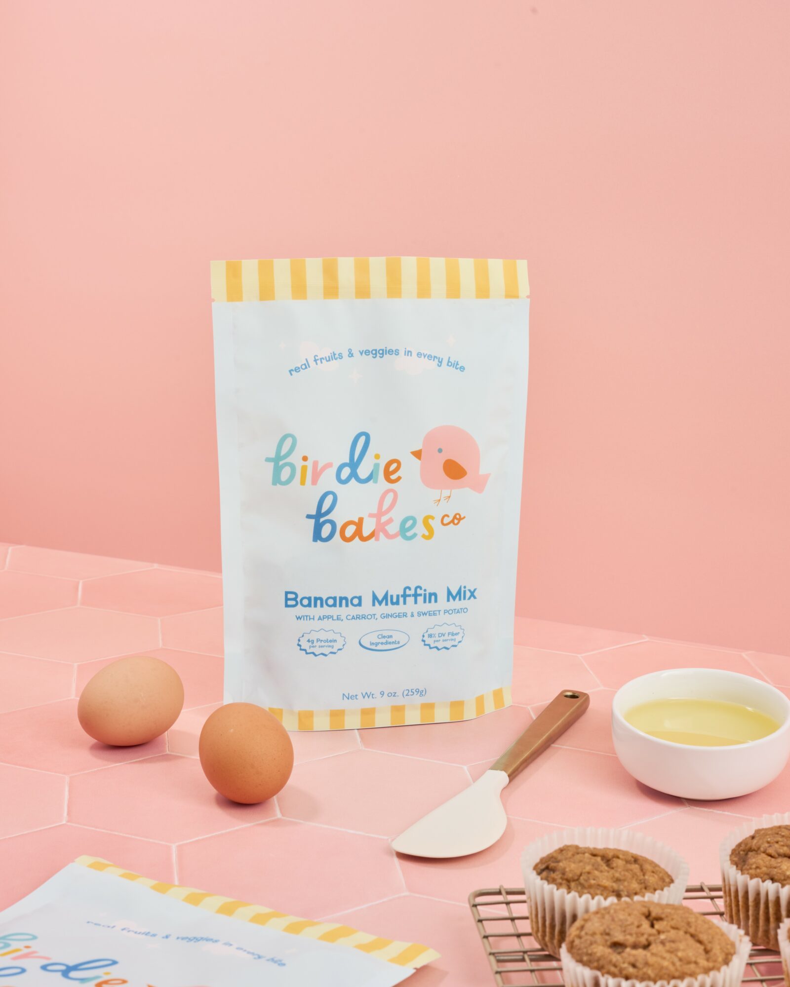 Banana muffin mix packet on pink surface with two eggs, a spatula, and muffins in paper cups.