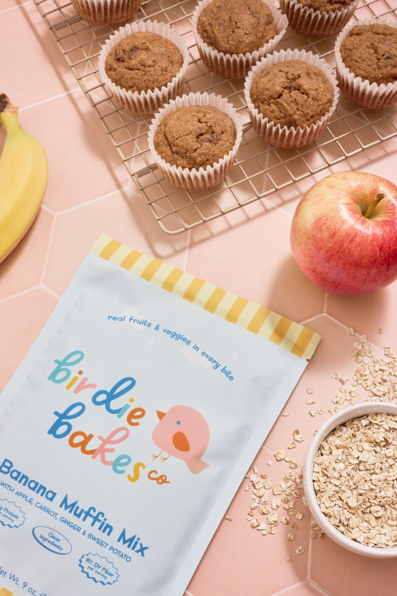 Banana muffins in paper cups on a cooling rack, apple, banana, and a bowl of oats on a pink surface, with a colorful booklet.
