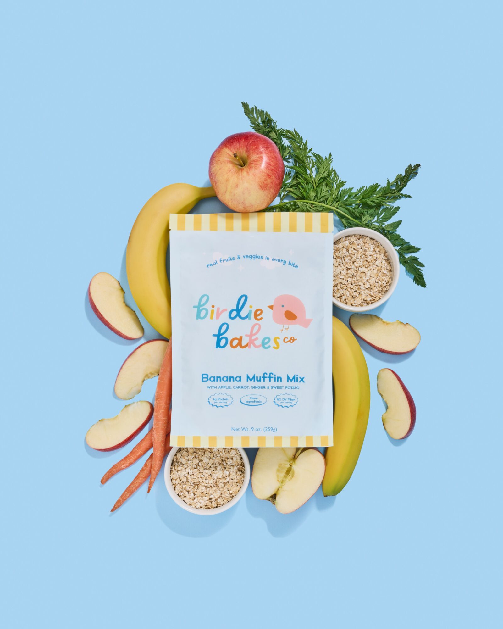 Banana, apple, carrots, leafy greens, and two bowls of muffin mix on a blue background.