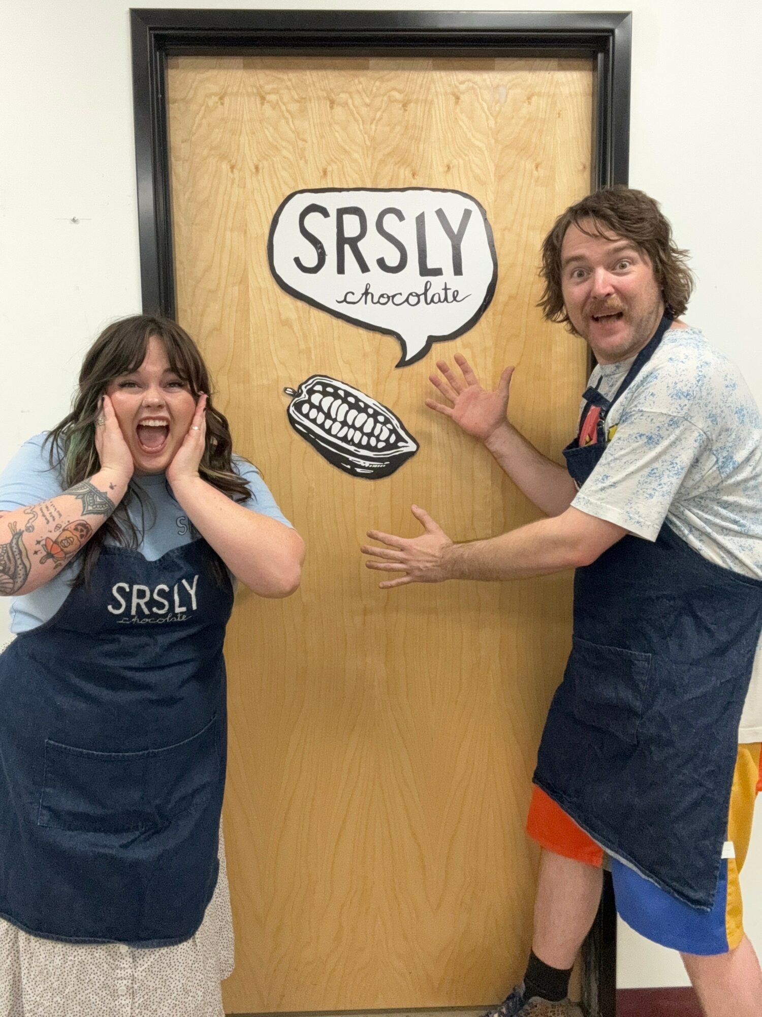 Two people smiling and gesturing in front of a door with a sign and drawings, one wearing an apron, the other in casual clothes.