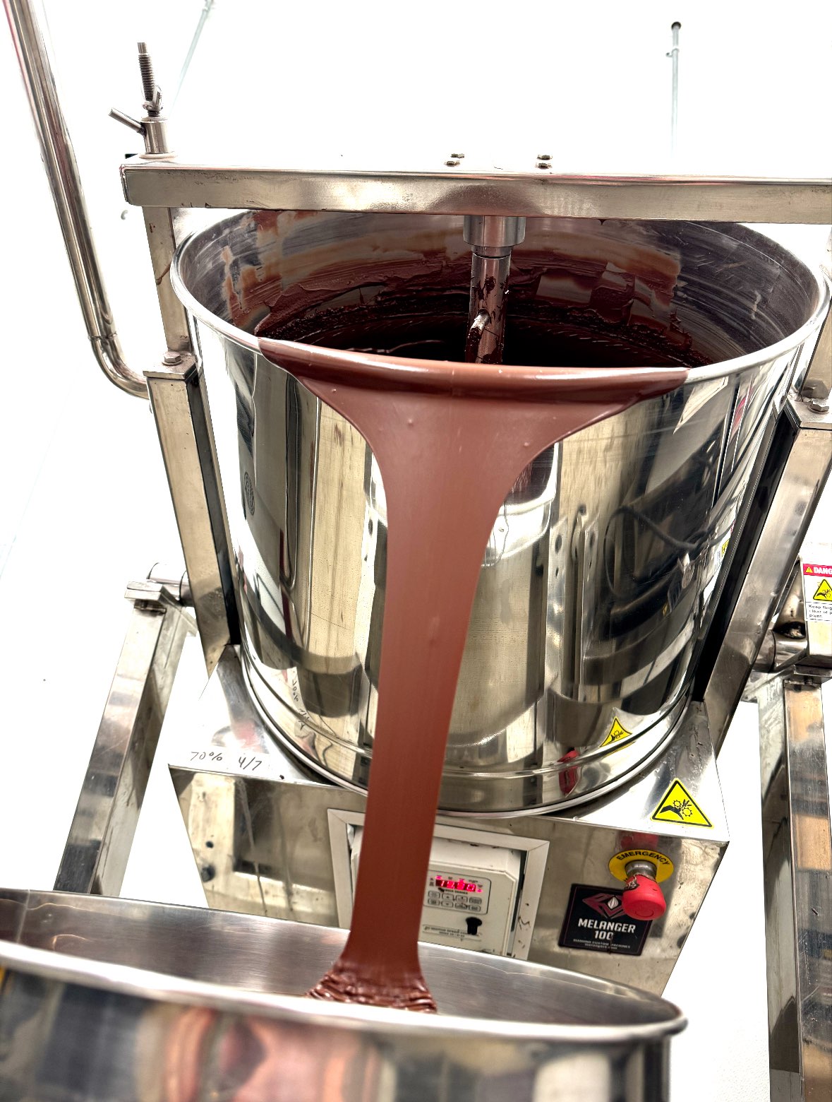 Chocolate liquid flows from a large stainless steel container into a tray.