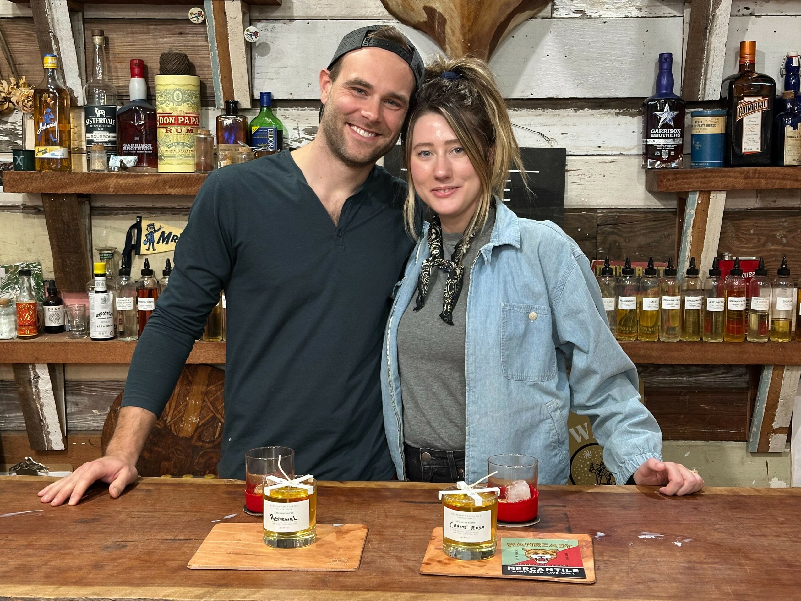 Two people standing behind a bar counter, smiling, with shelves of bottles in the background.