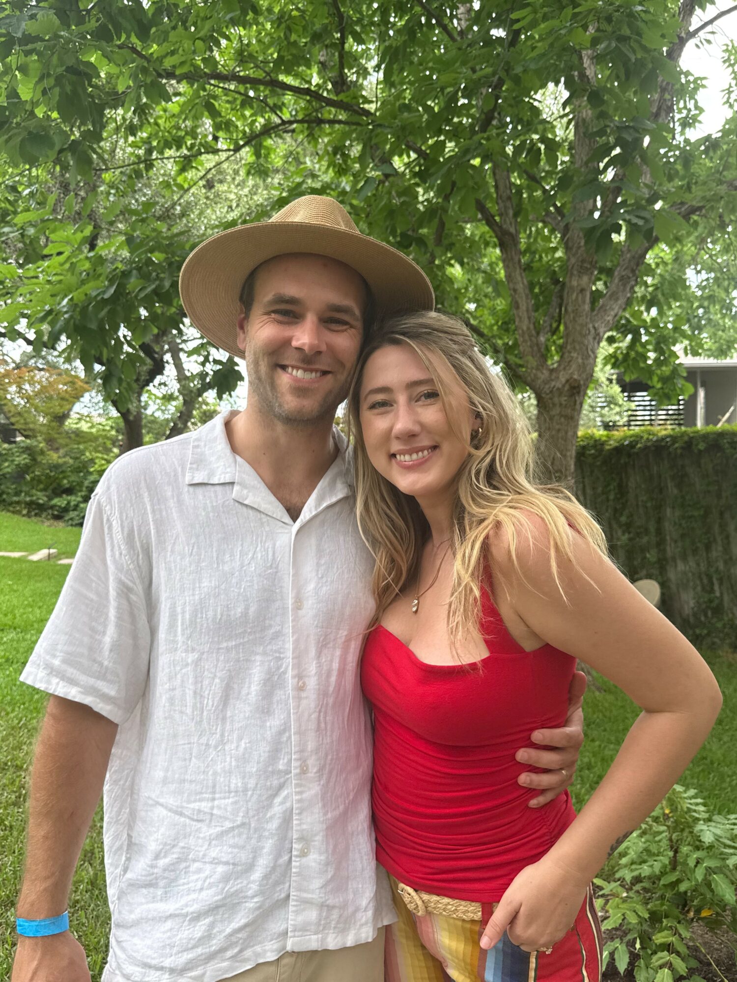 A smiling man and woman standing outdoors with trees and greenery in background.
