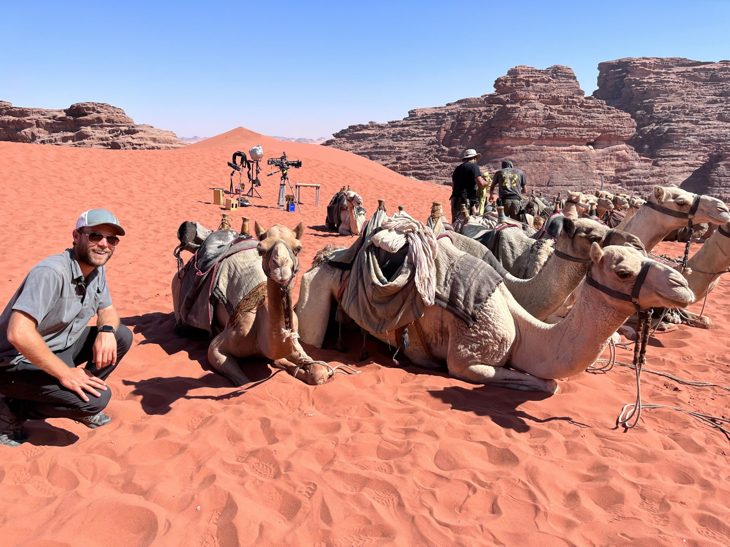 Person kneeling next to camels resting on red desert sand with rocky formations in background.