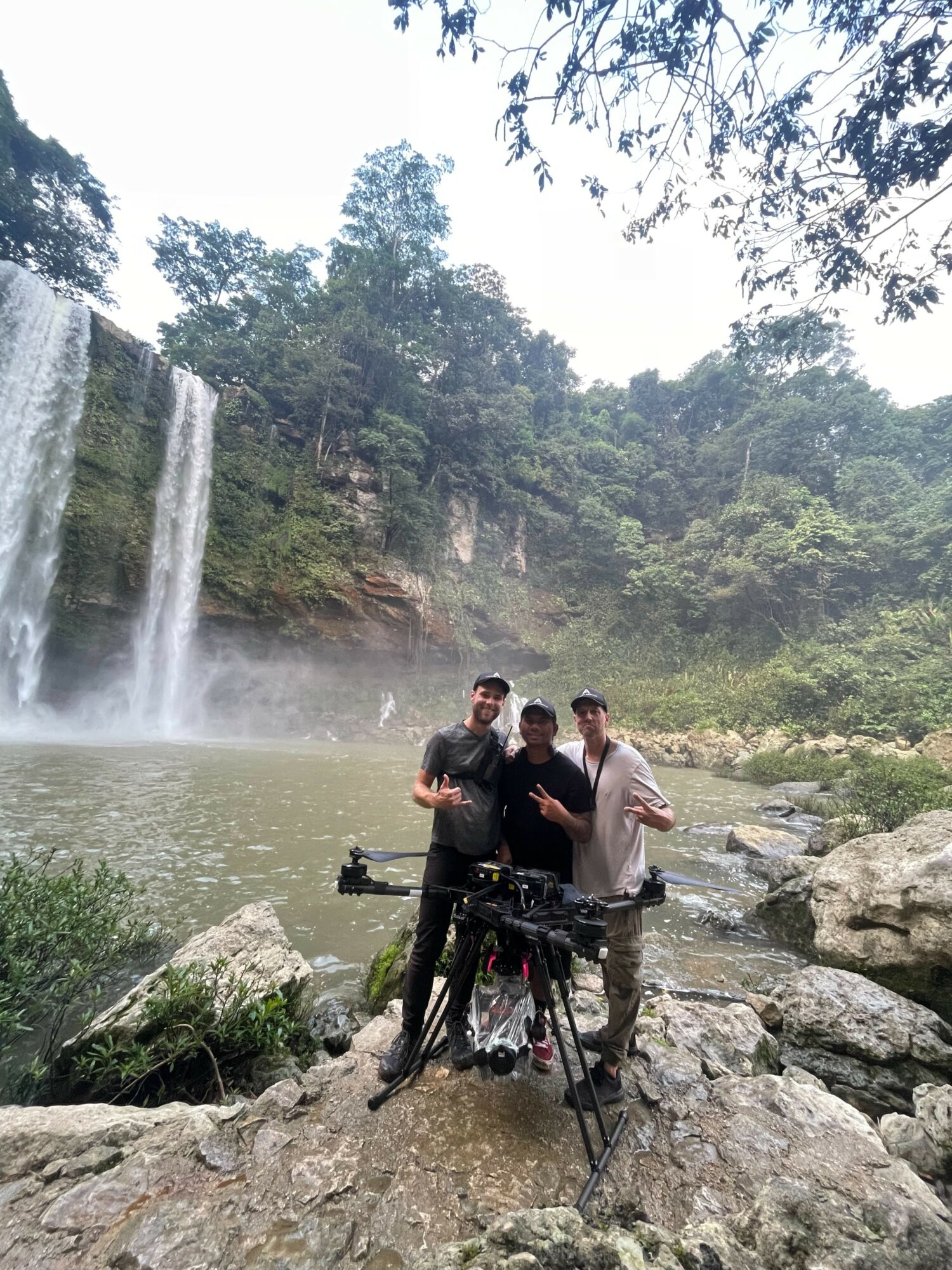 Three people standing on rocks near a waterfall and river, with trees and mist in the background.