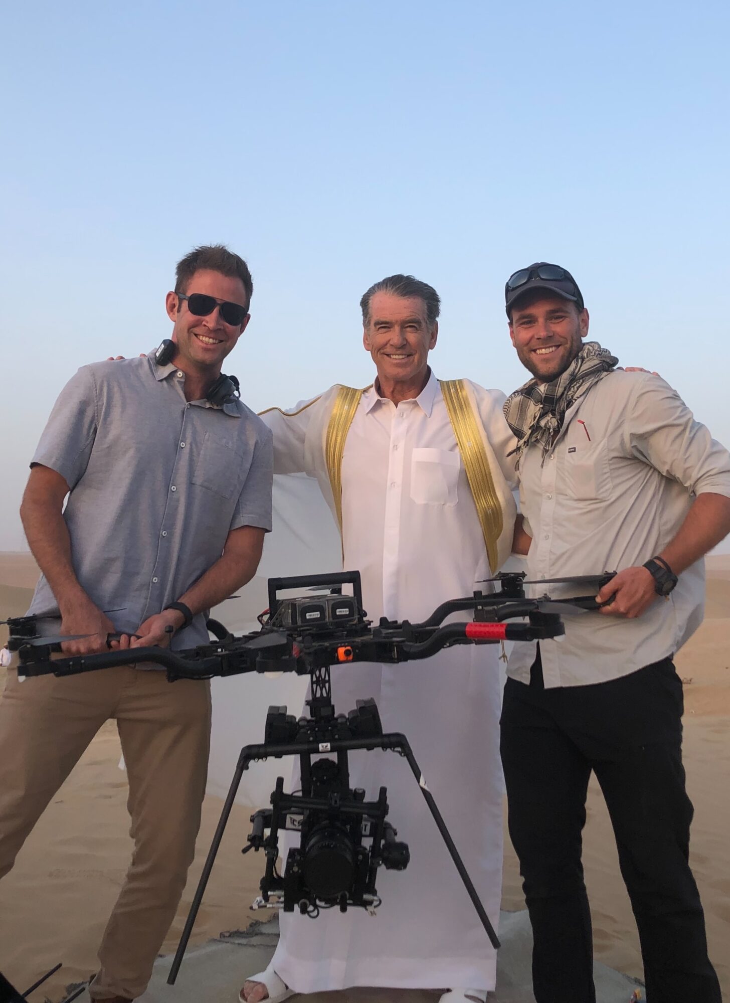 Three men standing outdoors with a camera mounted on a tripod, smiling, under a clear blue sky.