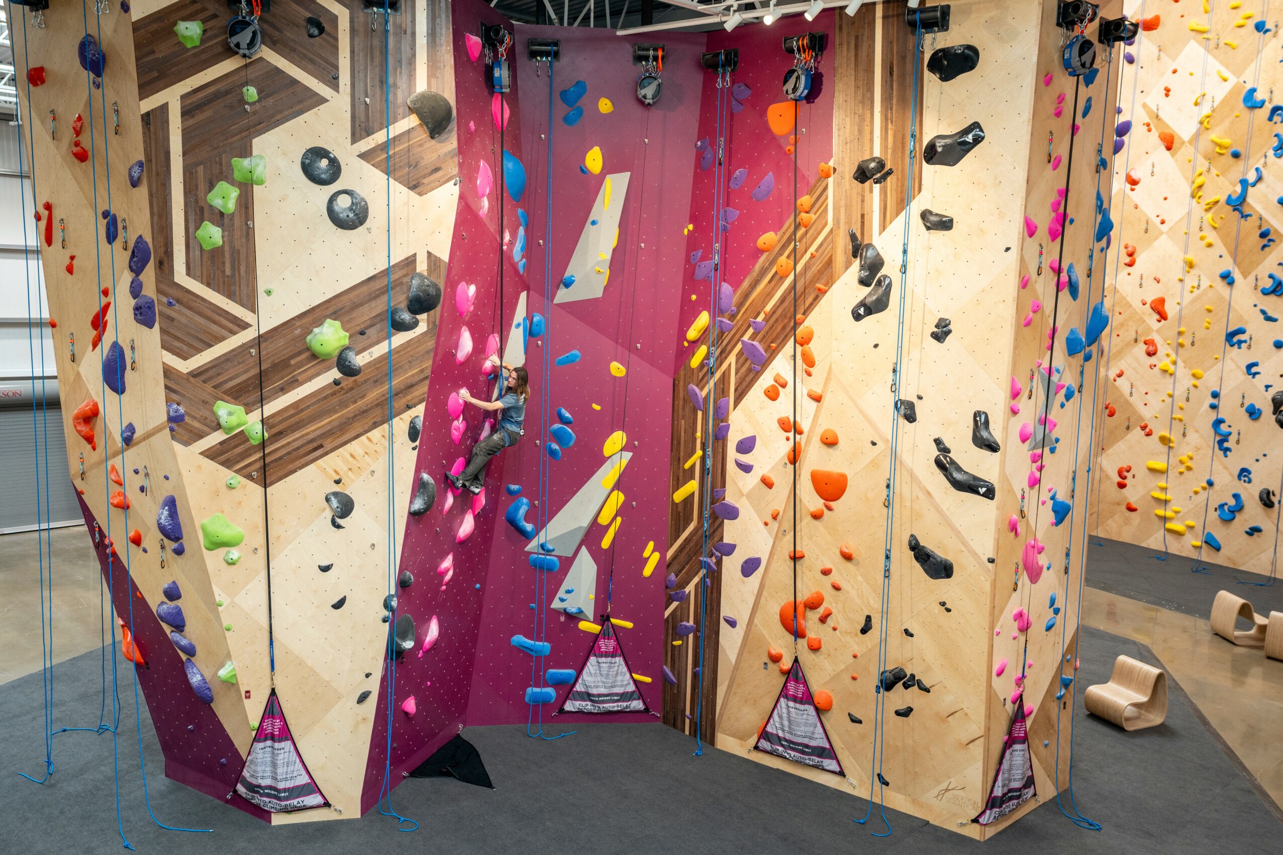 Indoor climbing wall with colorful holds and geometric patterns, surrounded by wooden panels and a blue floor.