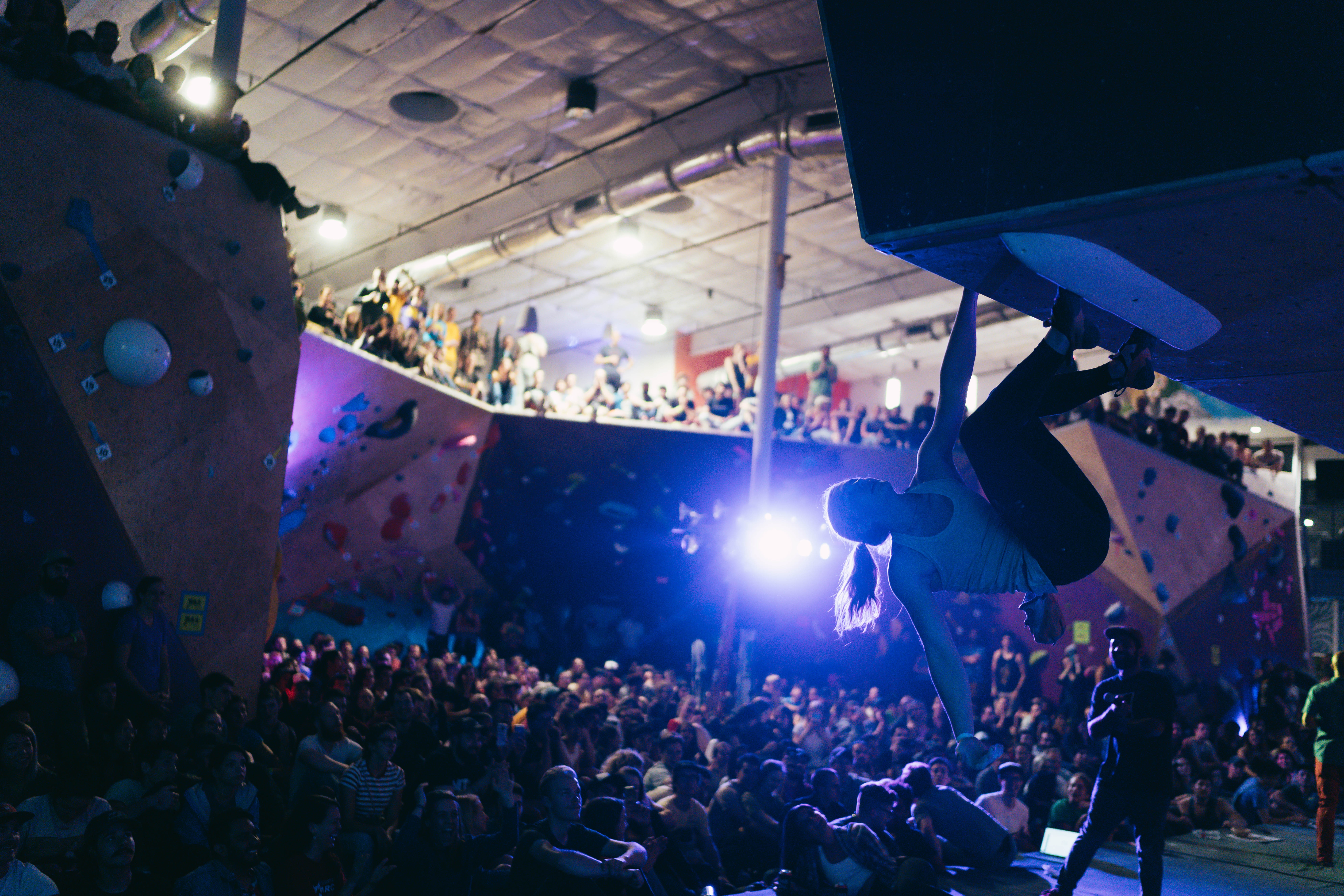 Indoor climbing gym with a crowd, person hanging upside down on climbing wall, bright lights, ceiling, and spectators.