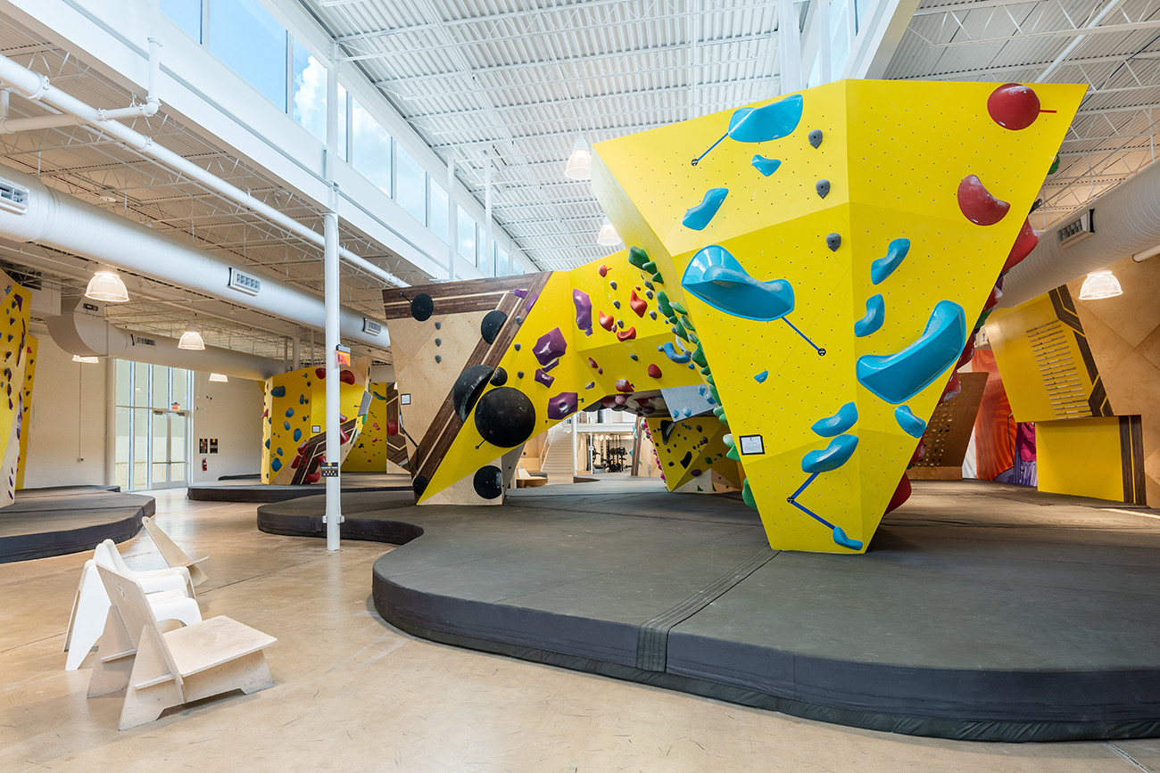 Indoor climbing gym with colorful rock climbing walls and padded floor, large windows, and seating area.