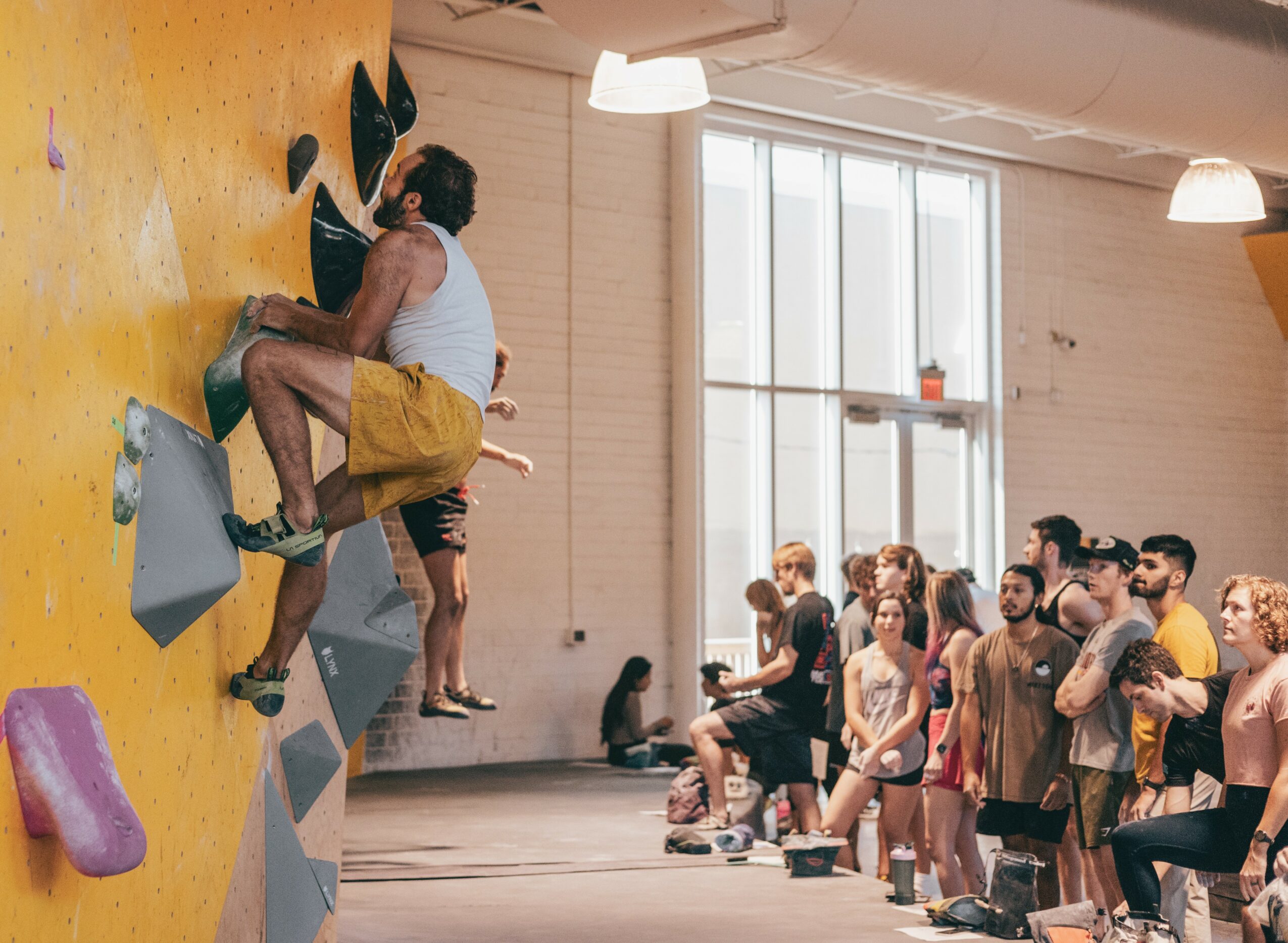 Person climbing indoor rock wall with crowd watching inside a spacious room with large windows.