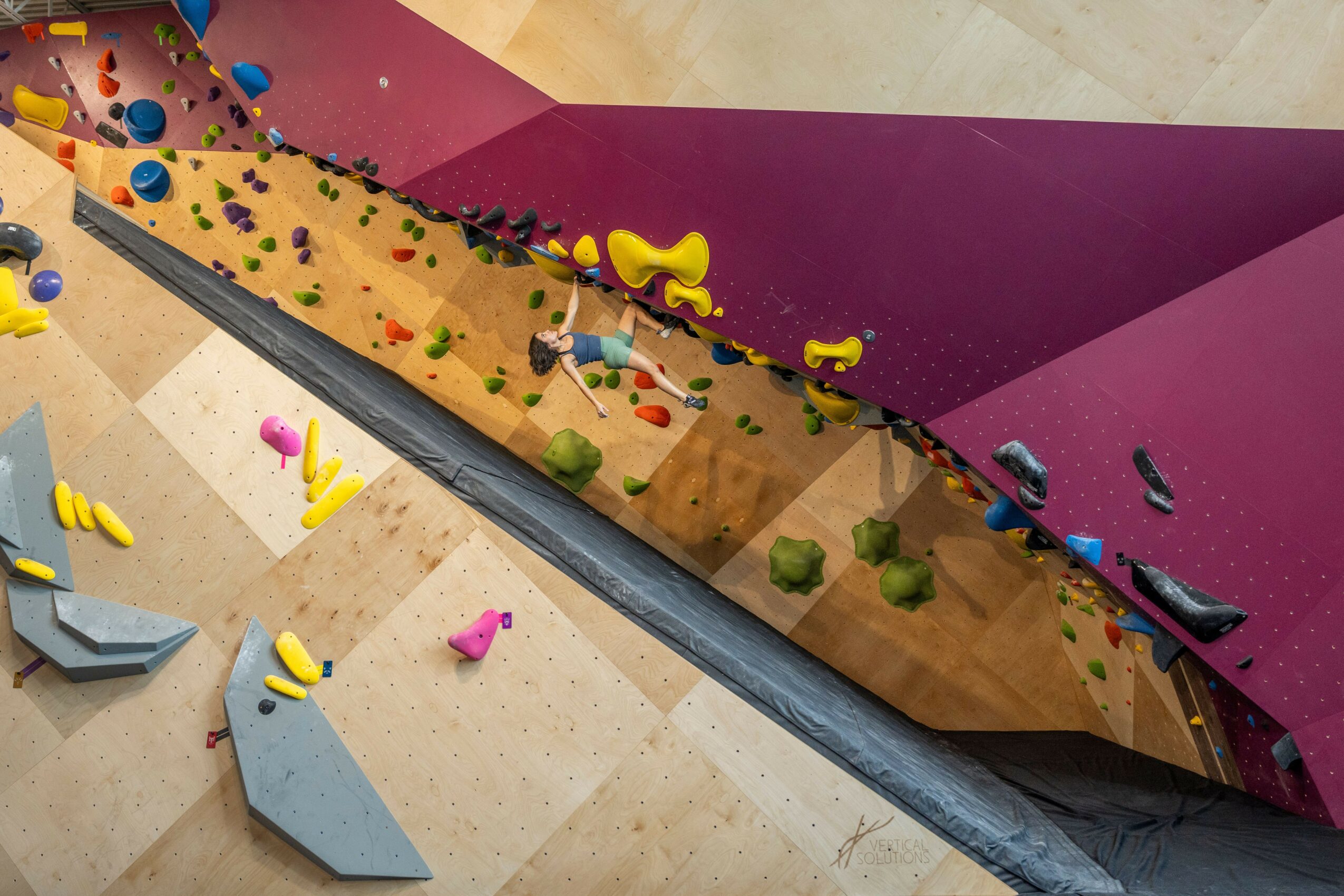 Indoor climbing wall with colorful holds and padded floor, featuring a steep incline and various climbing routes.