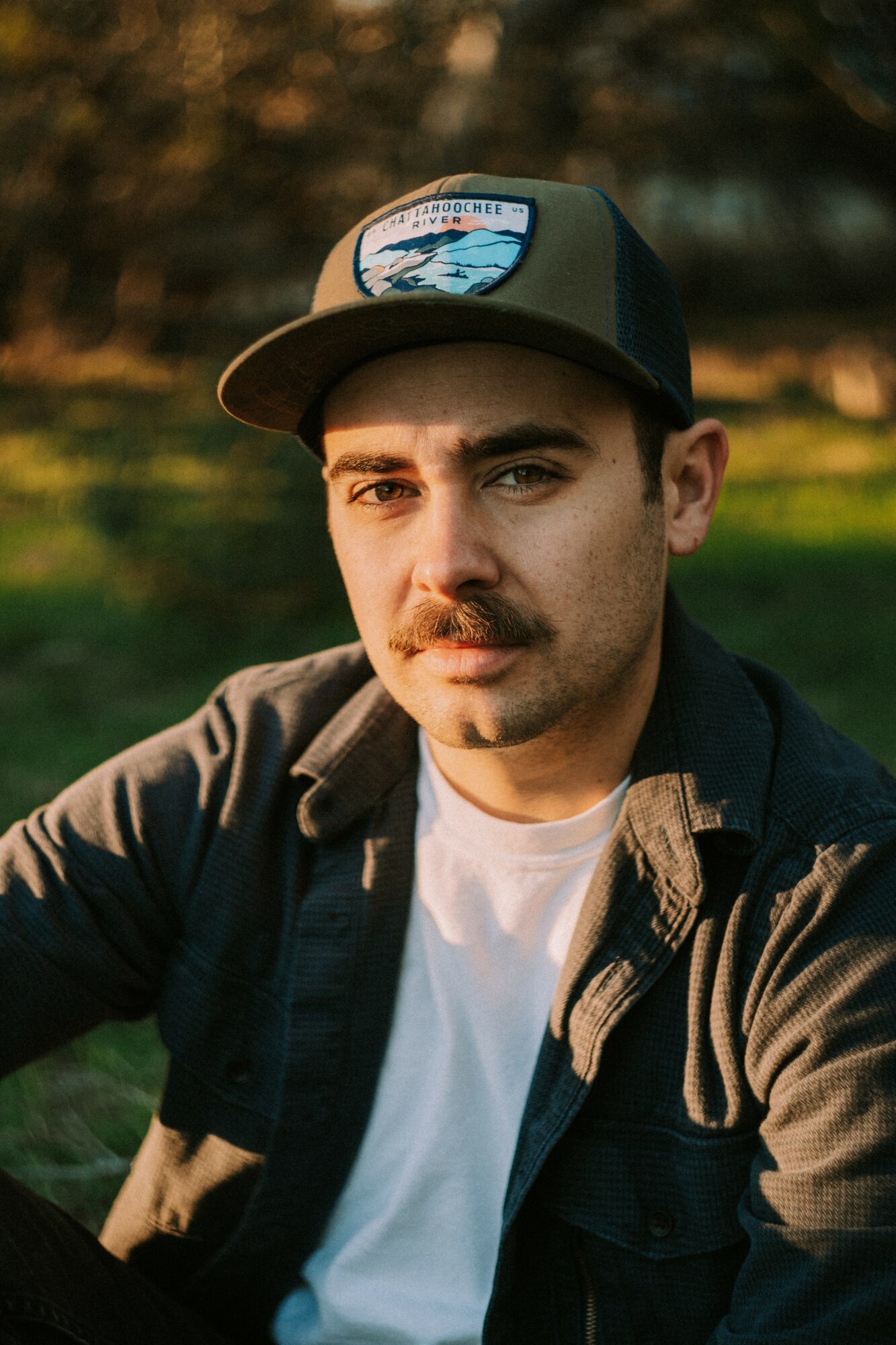 Young man with a mustache wearing a black cap and jacket outdoors, looking at the camera.