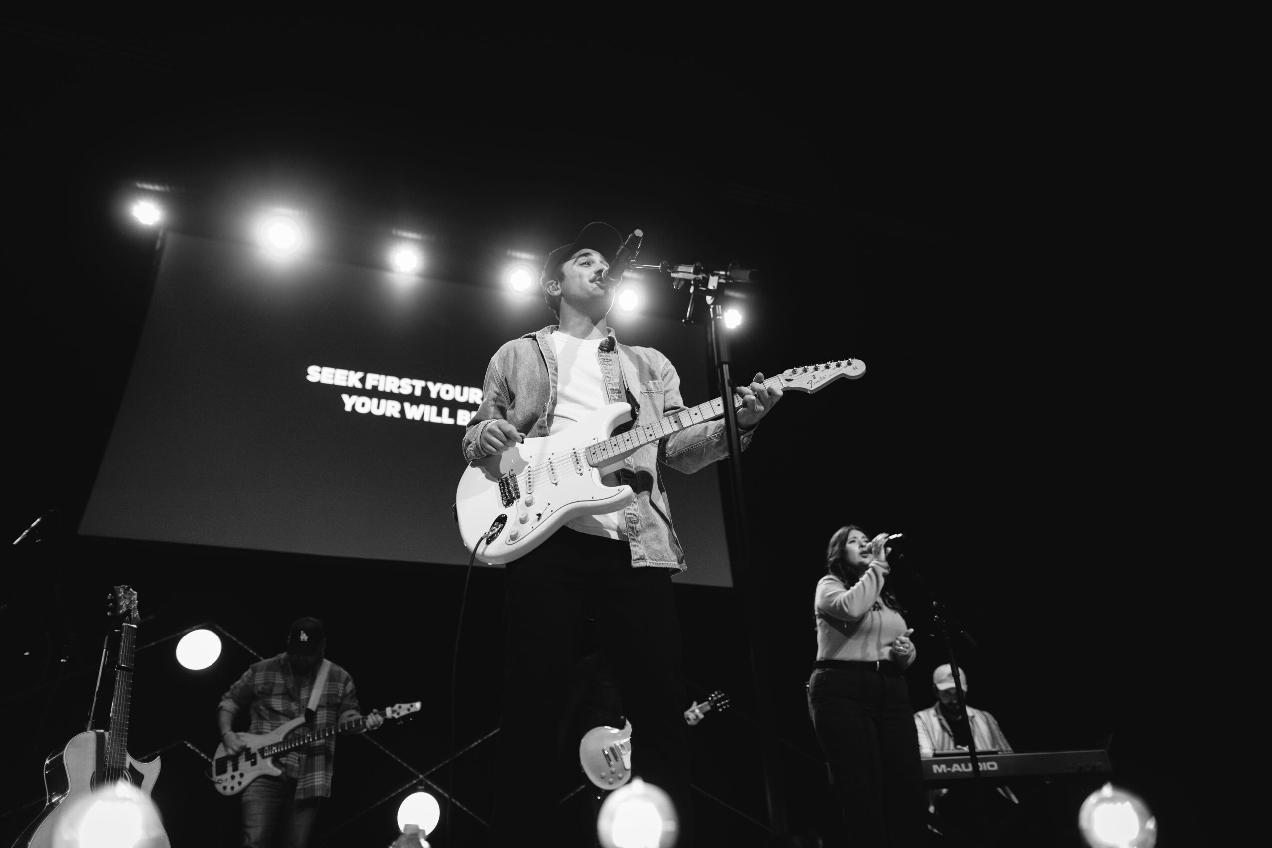 Musician playing electric guitar on stage with band members, bright stage lights, and a large screen in the background.