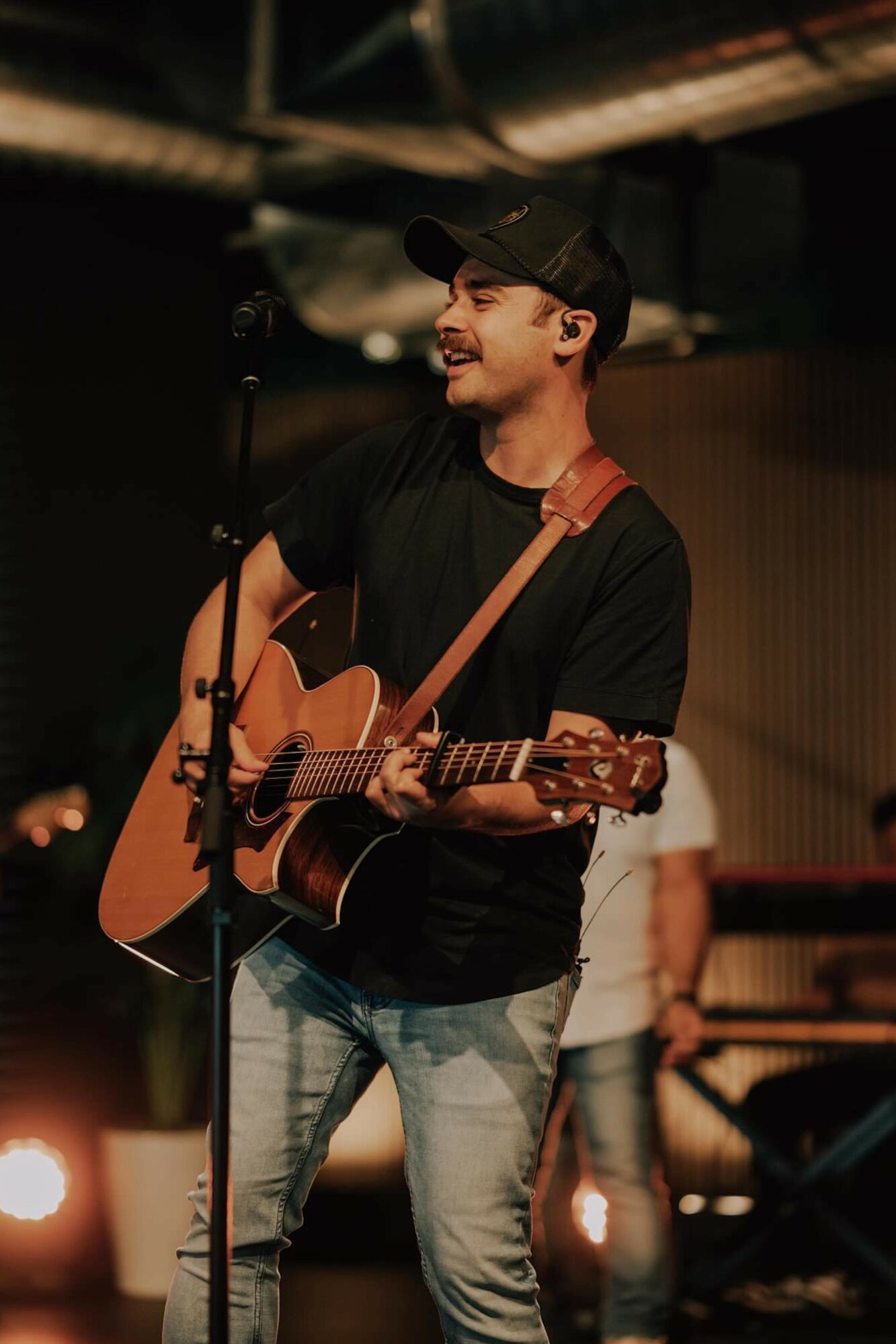 Young man playing acoustic guitar on stage, smiling, wearing a black cap and t-shirt, with a dark background.