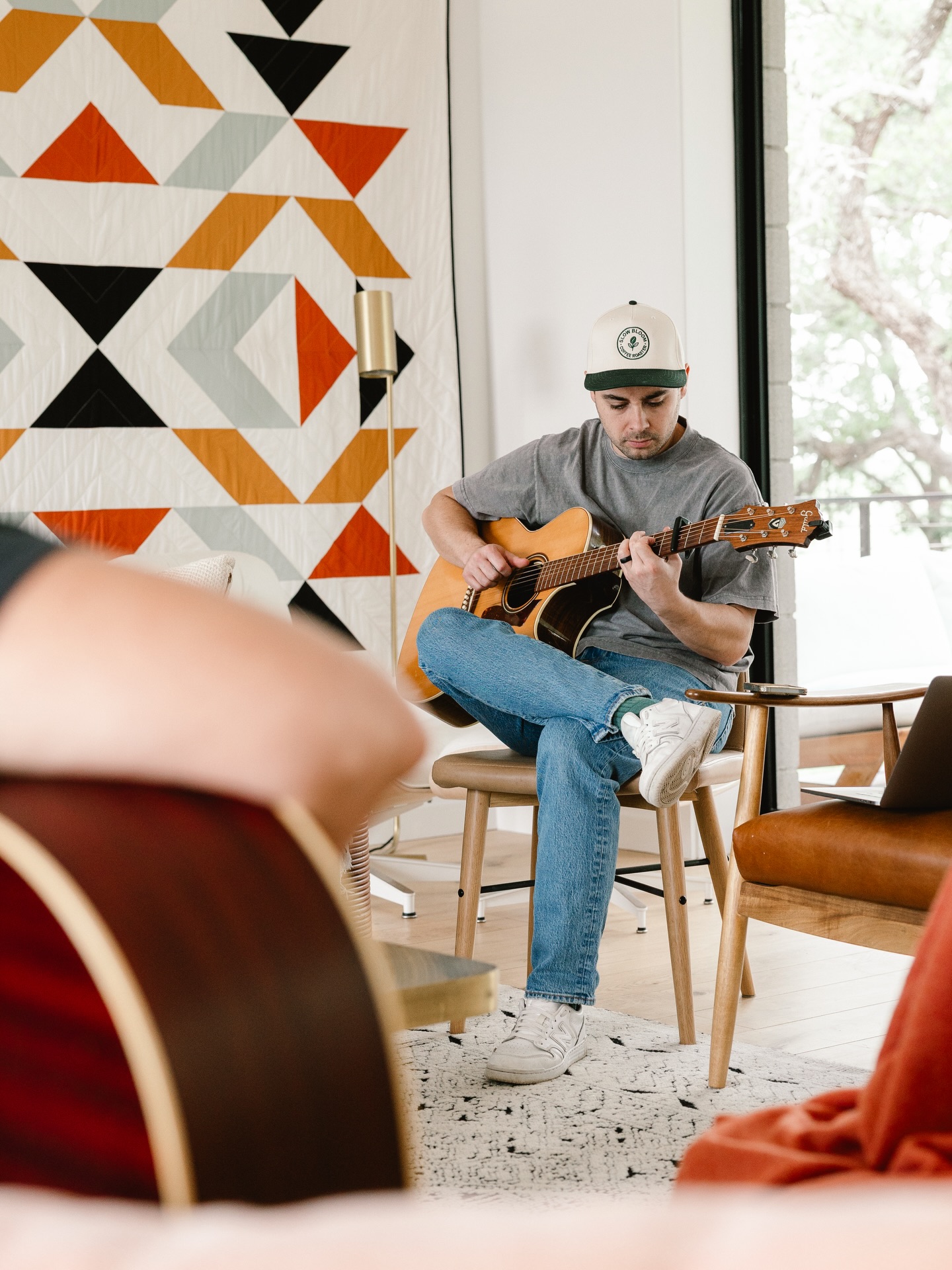 Young man playing acoustic guitar indoors, sitting on a chair, wearing a cap and casual clothes, with a geometric wall pattern behind him.