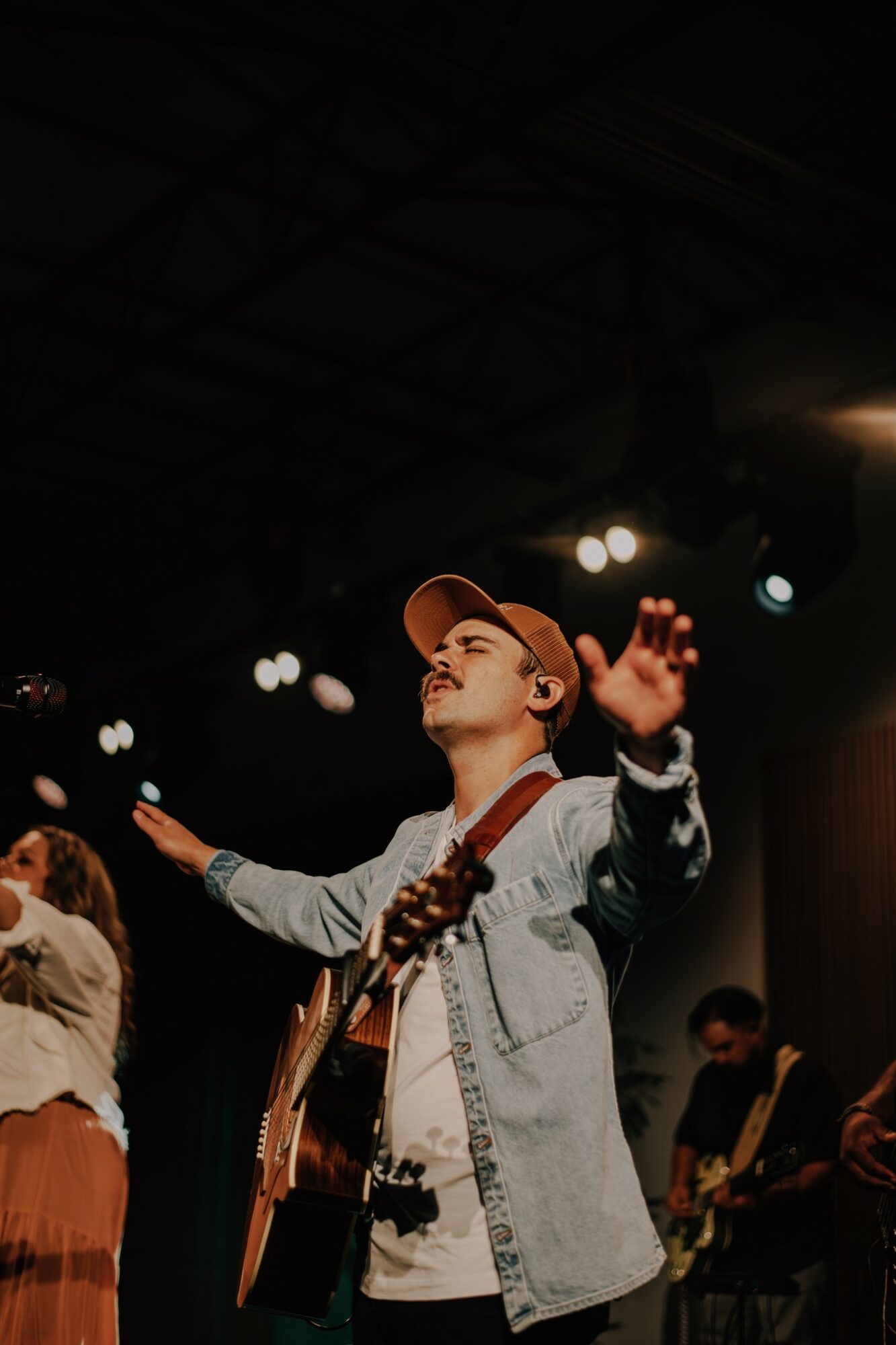 Young person with a hat playing guitar on stage with arms raised, stage lights in background.