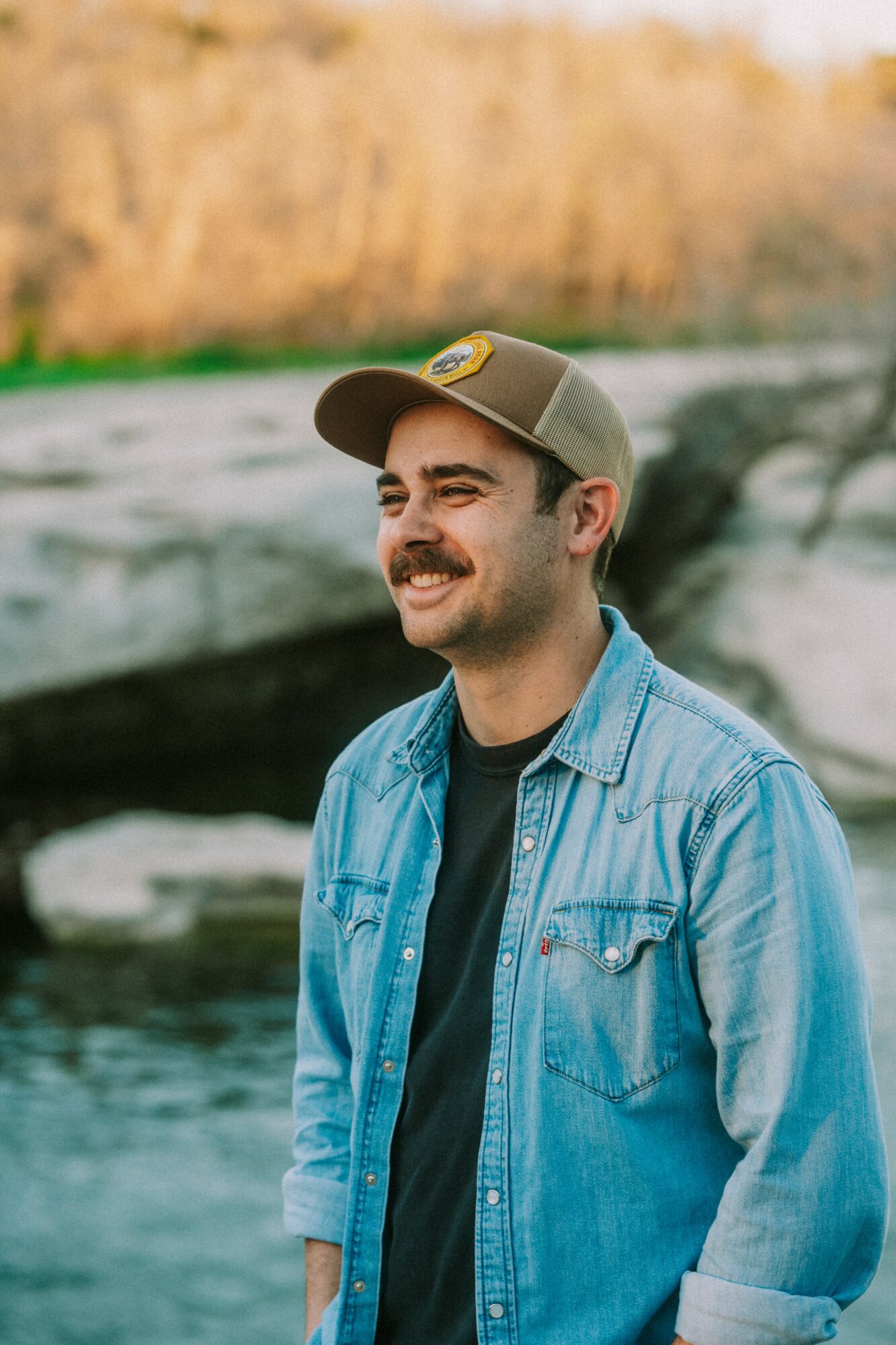 Man with a mustache wearing a baseball cap and denim jacket outdoors near water and rocks.