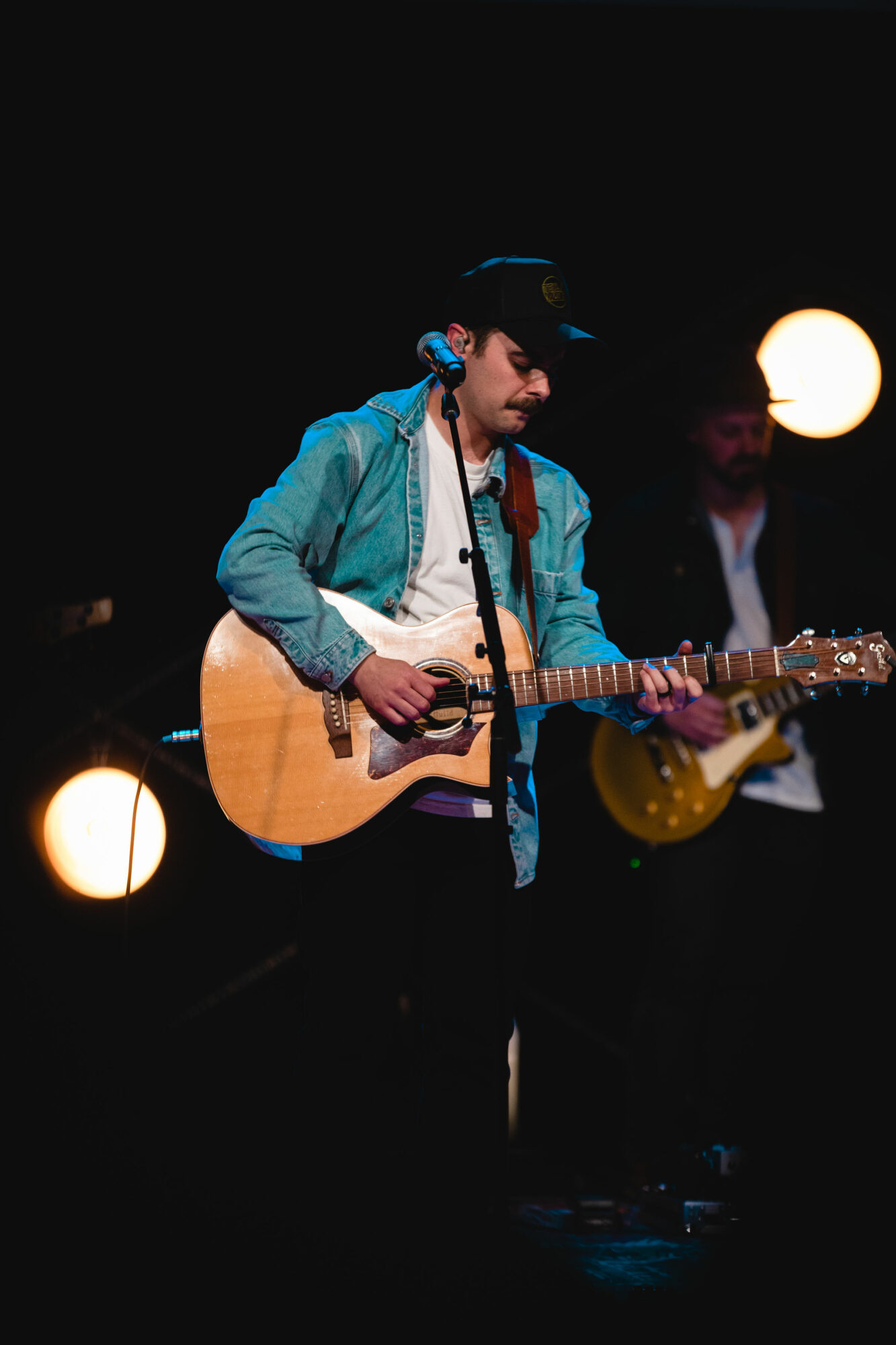 Young man playing acoustic guitar on stage with a person in the background, illuminated by stage lights.