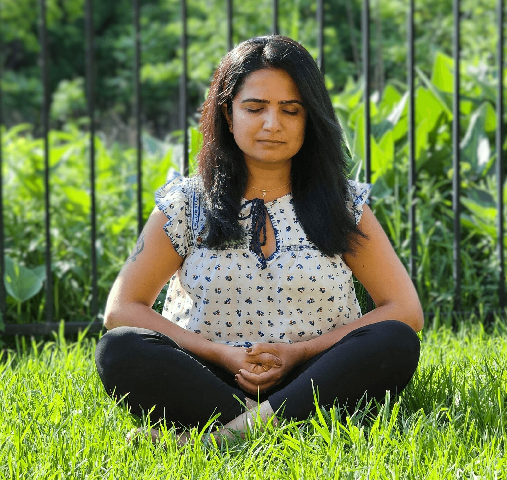 Woman sitting cross-legged on grass with eyes closed, outdoors with green plants and a fence behind her.