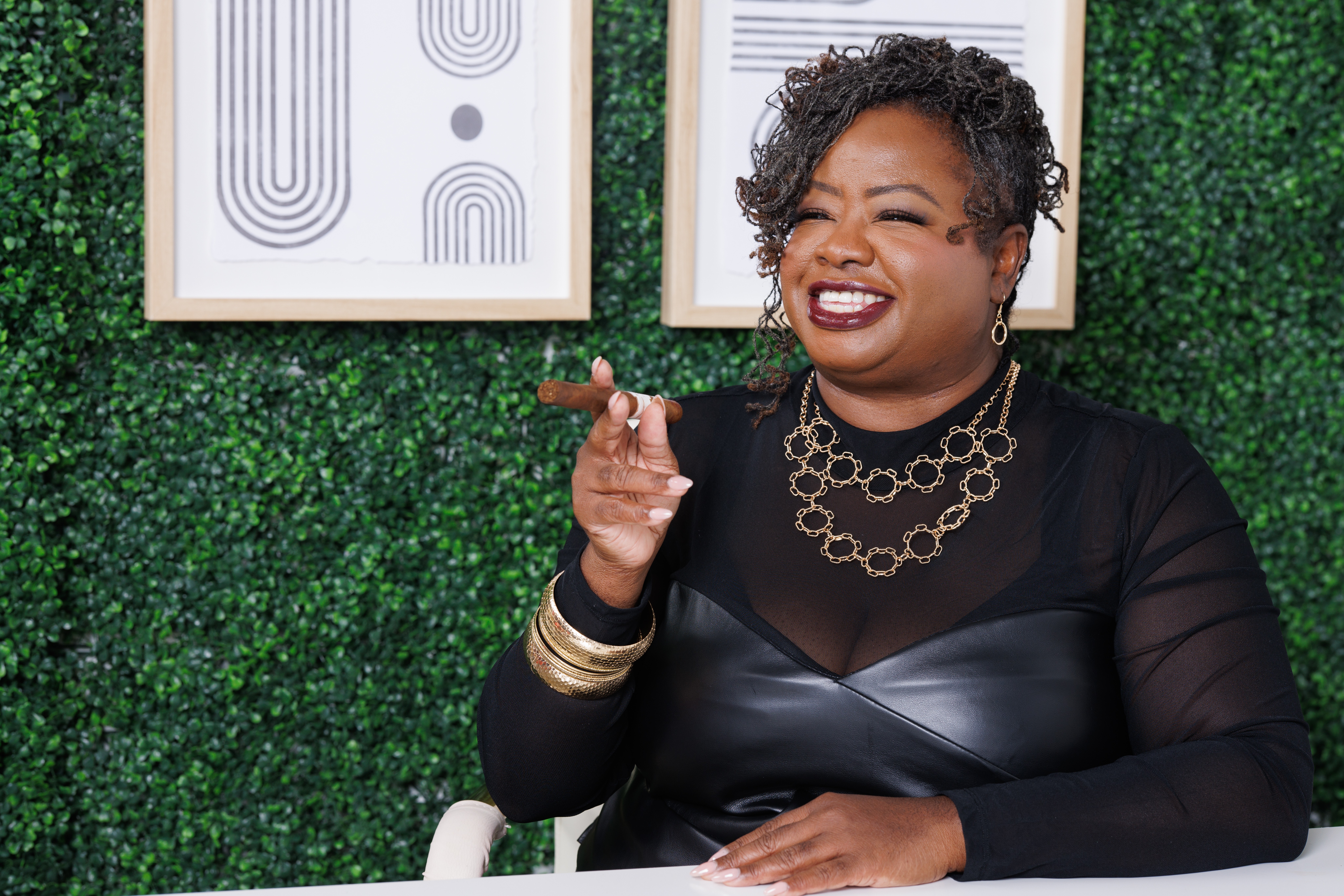 Smiling woman with short curly hair, wearing black top and gold jewelry, sitting at a table with green wall background.